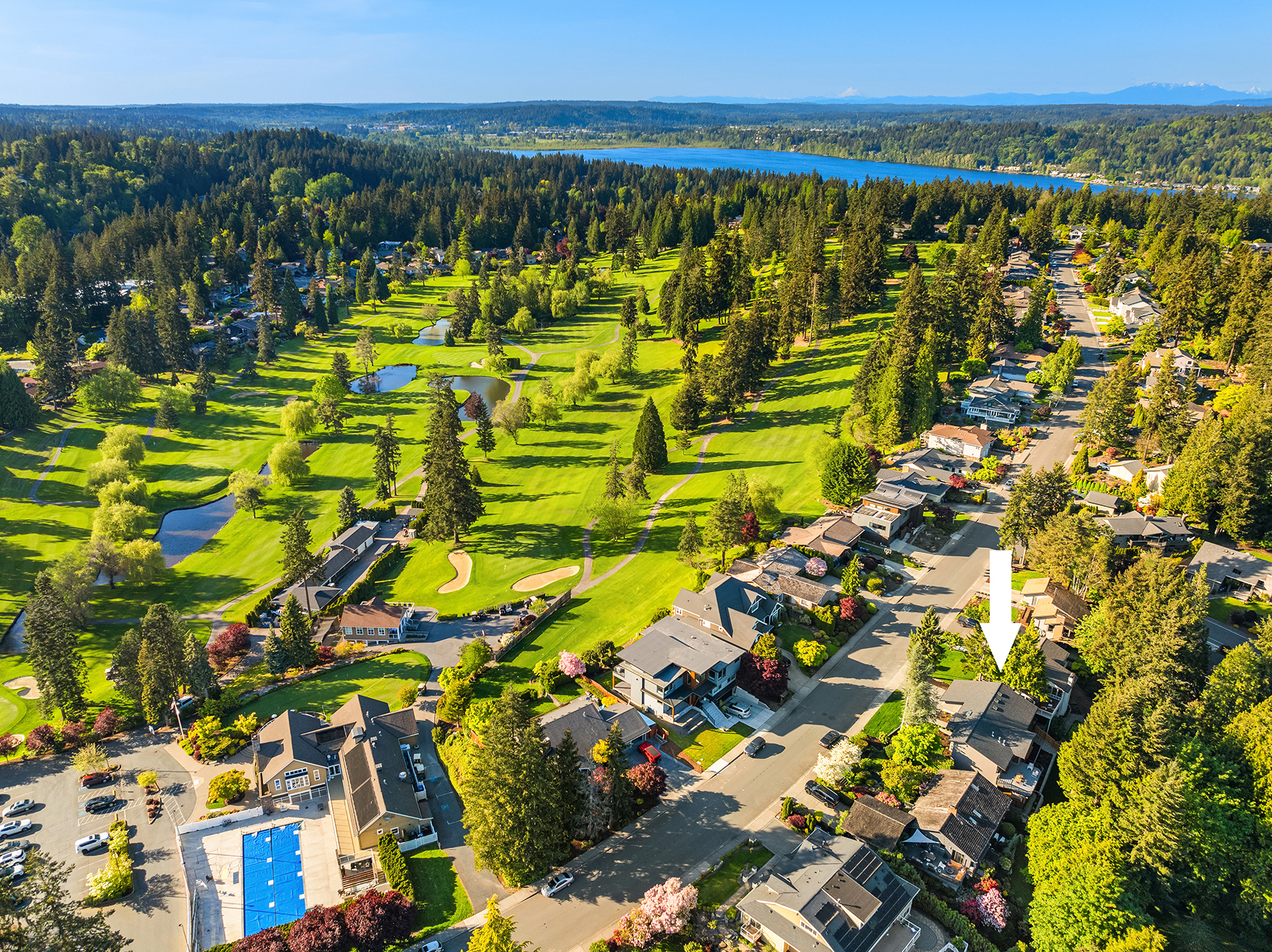 Aerial view of a suburban neighborhood next to a green golf course with fairways, trees, and ponds; a white arrow points to a specific house. Blue lake and forested hills visible in the background.