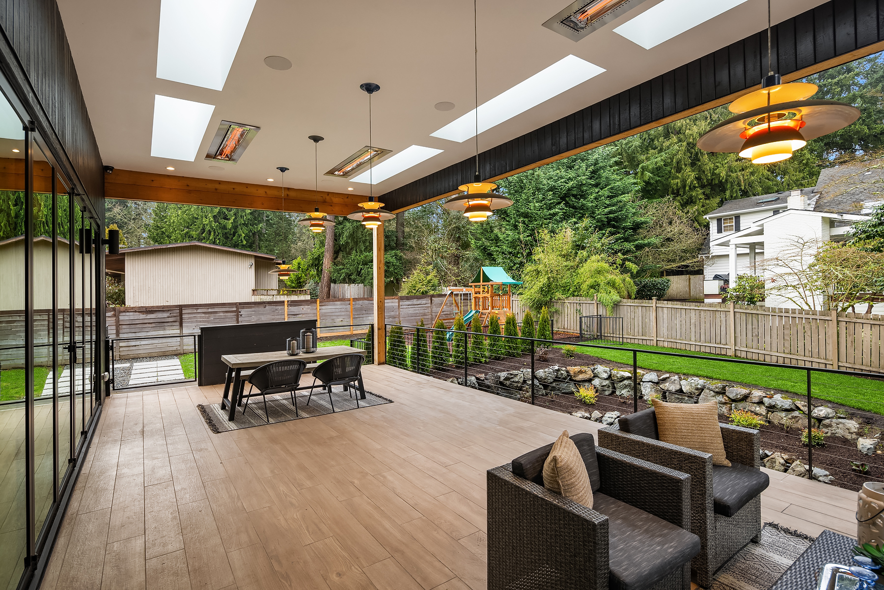 Covered outdoor patio with modern seating and dining areas, ceiling heaters, skylights, and view of a backyard with a childrens playset, green lawn, and neighboring houses surrounded by trees and fencing.
