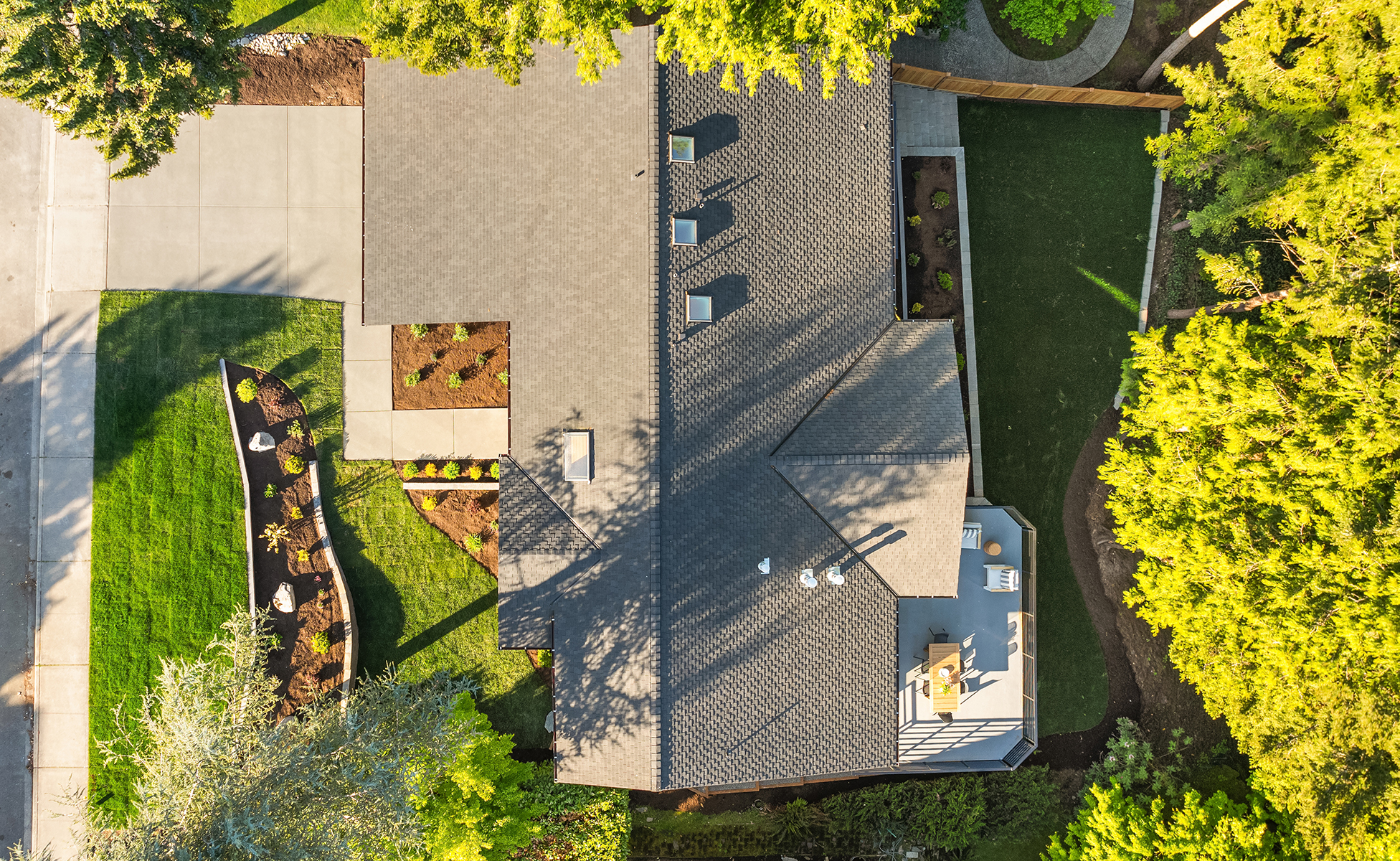 Aerial view of a house with a gray roof, driveway, green lawn, landscaped garden beds, and surrounding trees casting shadows. A patio with outdoor furniture is visible on one side of the house.
