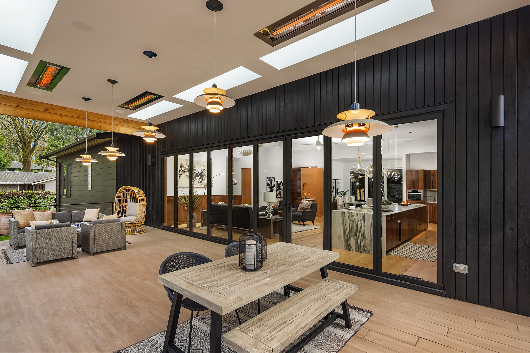 Modern outdoor patio with a wooden dining table, black chairs, hanging lights, and lounge area. Large glass doors reveal a stylish interior with contemporary decor and wood accents.