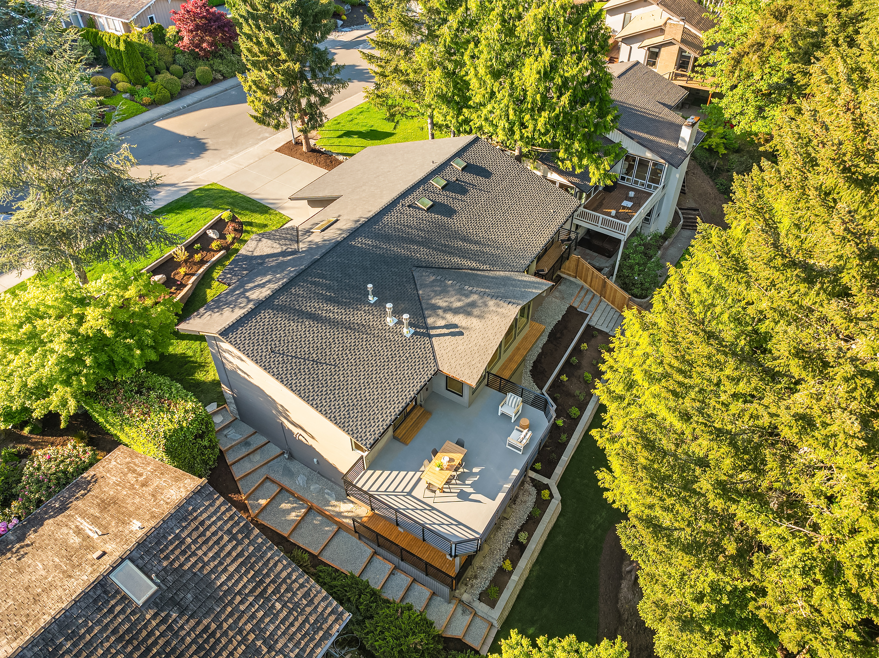 Aerial view of a modern house with a gray roof, spacious deck with outdoor furniture, landscaped yard, and surrounding trees and neighboring homes on a sunny day.