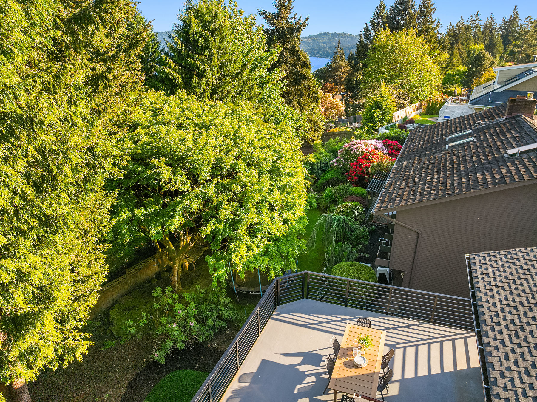 A sunny backyard with lush green trees and bushes, seen from above. A wooden deck features a table and chairs, while nearby houses and distant mountains are visible under a clear blue sky.