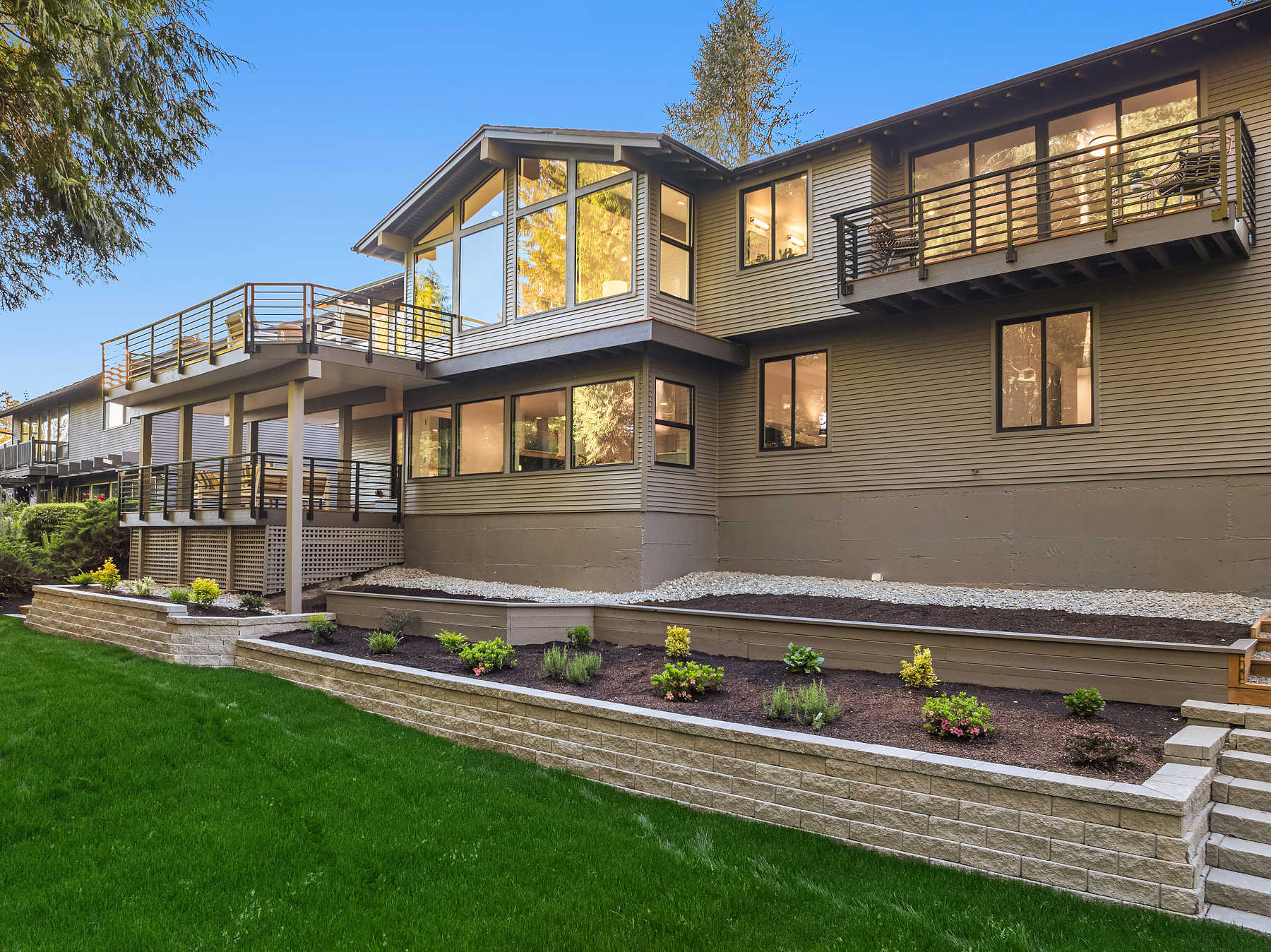 Modern two-story house with large windows, multiple balconies with metal railings, and a well-maintained yard featuring green grass, stone retaining walls, and freshly planted shrubs and flowers.
