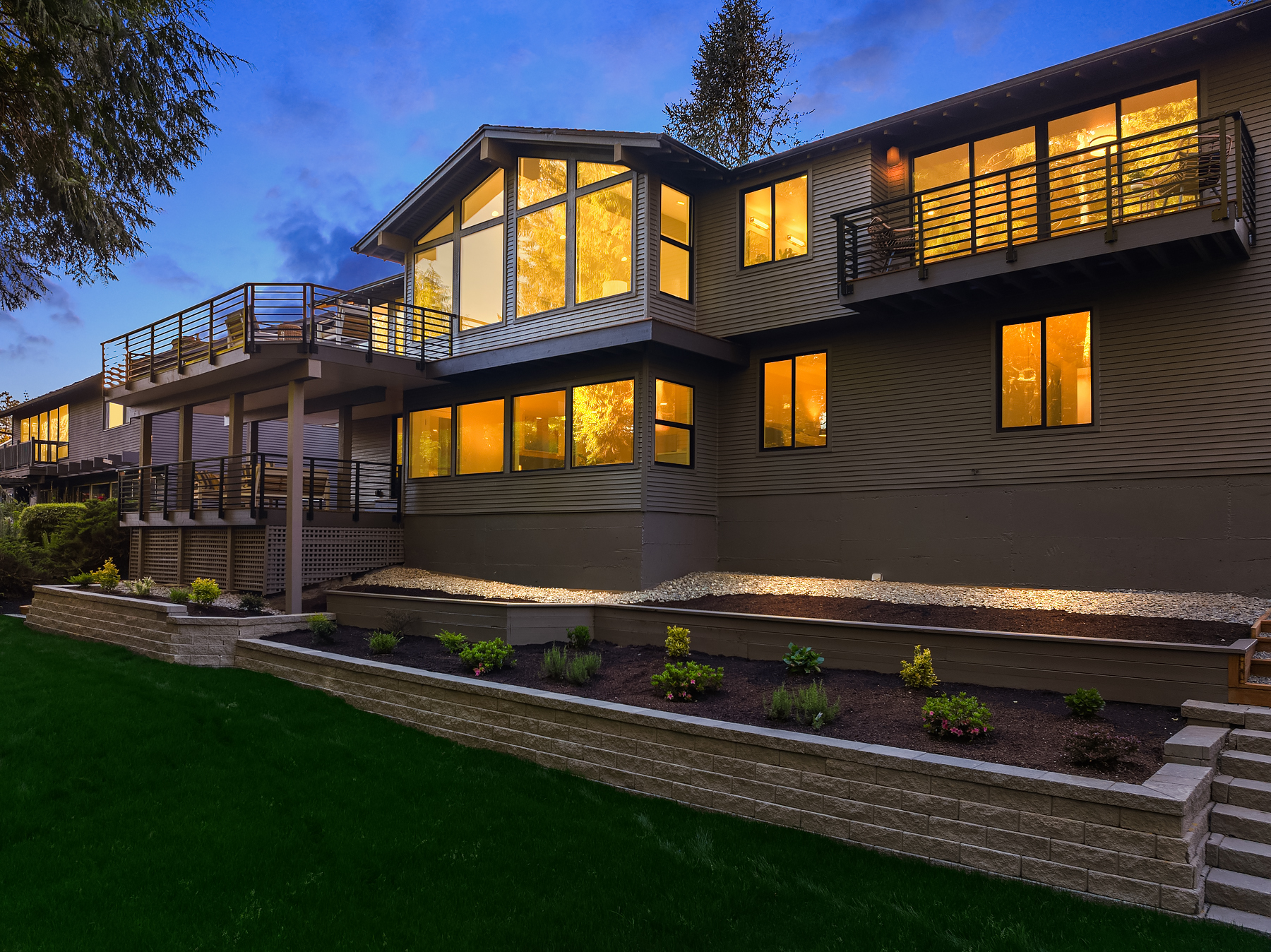 A modern two-story house at dusk with large windows glowing with yellow light, multiple balconies with railings, and a landscaped garden with retaining walls and green grass in the foreground.