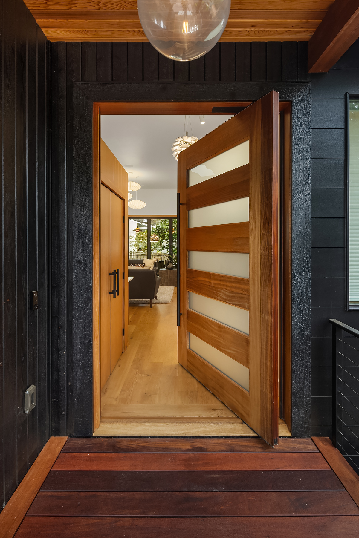 A modern wooden front door with frosted glass panels is partially open, revealing a bright interior hallway with light wood floors and a view into a living room and backyard.