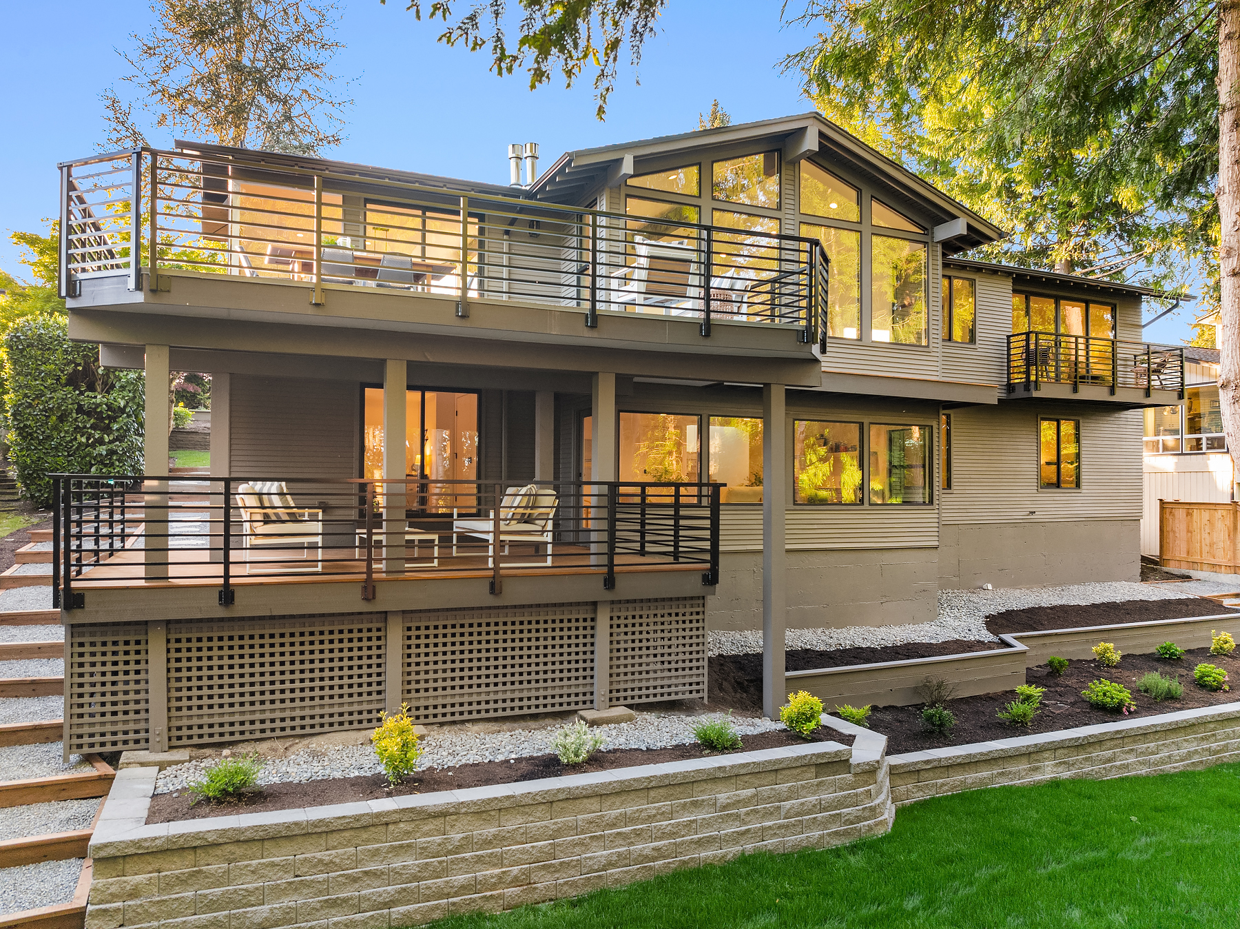Modern two-story house with large windows, multiple balconies with black railings, outdoor seating area, and landscaped front yard with green grass and small shrubs.