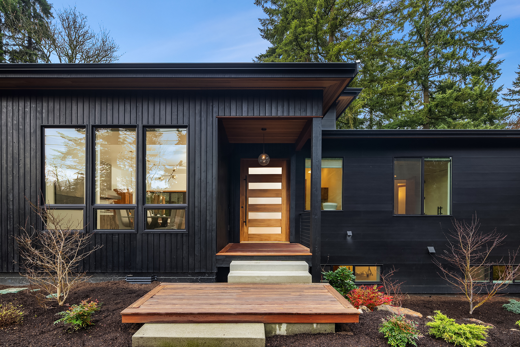 Modern black house with large windows, a wooden front door with horizontal glass panels, a small porch, and minimal landscaping in the front yard, surrounded by tall evergreen trees under a blue sky.