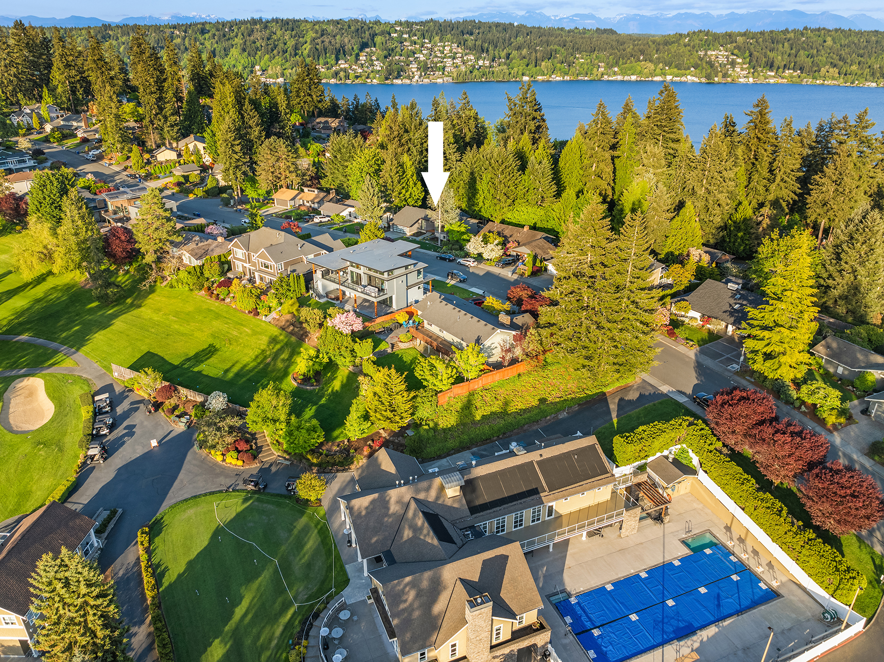 Aerial view of a suburban neighborhood near a lake with a golf course, clubhouse, and swimming pool in the foreground. An arrow points to a specific two-story house surrounded by trees and other homes.