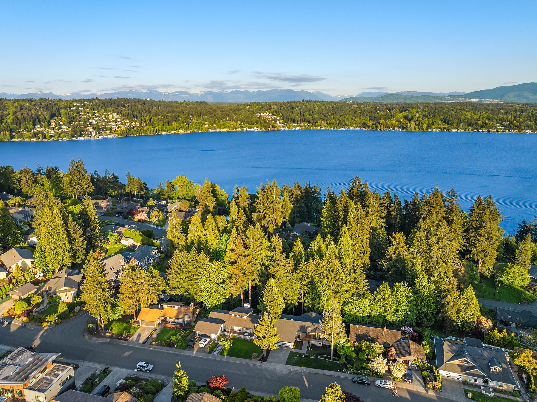 Aerial view of a suburban neighborhood with houses and tree-lined streets beside a large blue lake, with forested hills and distant mountains under a clear blue sky.