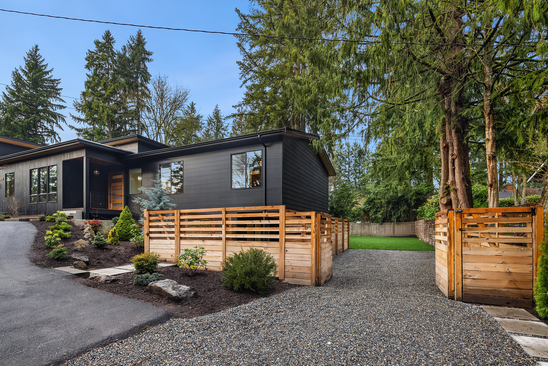 Modern black house with large windows, surrounded by tall trees and landscaped plants. A wooden fence and gate enclose a gravel driveway, leading to a lush green backyard. The sky is clear and the setting feels peaceful.