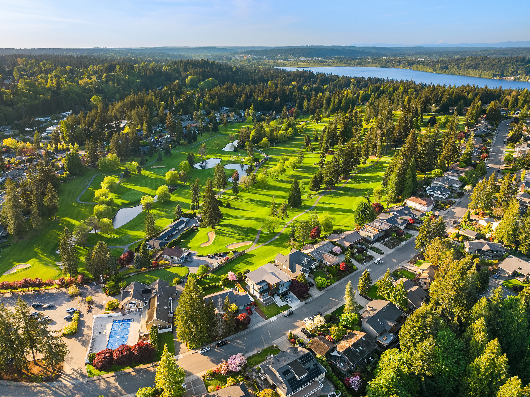 Aerial view of a suburban neighborhood bordering a golf course with green fairways, sand traps, trees, and a lake in the background under a clear blue sky.