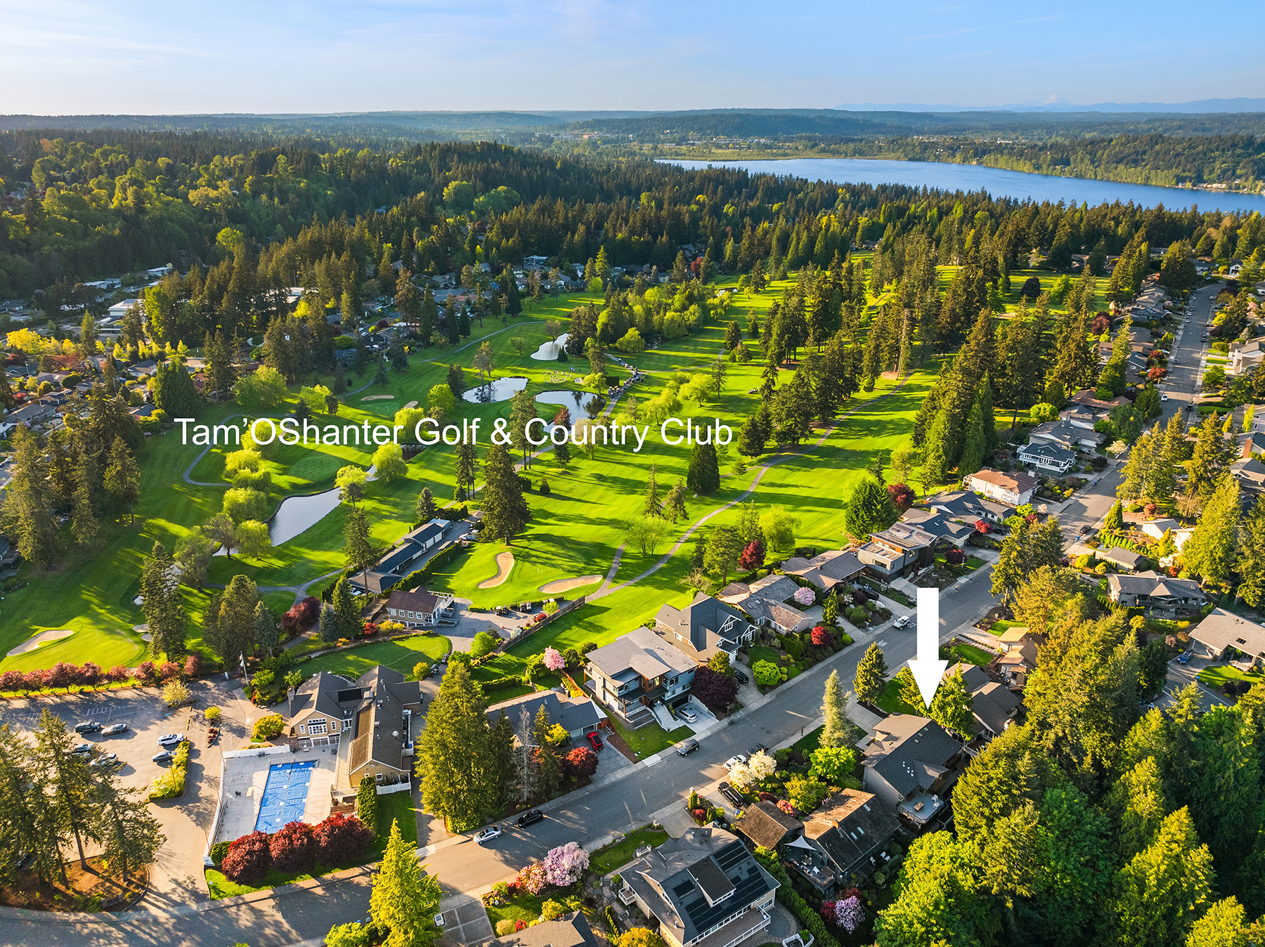 Aerial view of a green golf course with surrounding trees and houses. A white arrow points to a specific house near the golf course, labeled as Tam O’Shanter Golf & Country Club. A pool and winding roads are also visible.