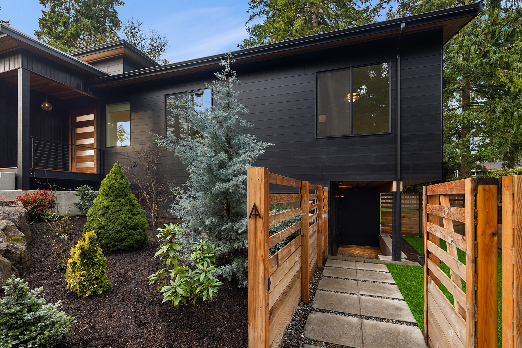 Modern house with dark exterior, large windows, and wooden accents. A wooden fence lines a stone walkway leading to the entrance, surrounded by neatly landscaped greenery and trees under a clear sky.