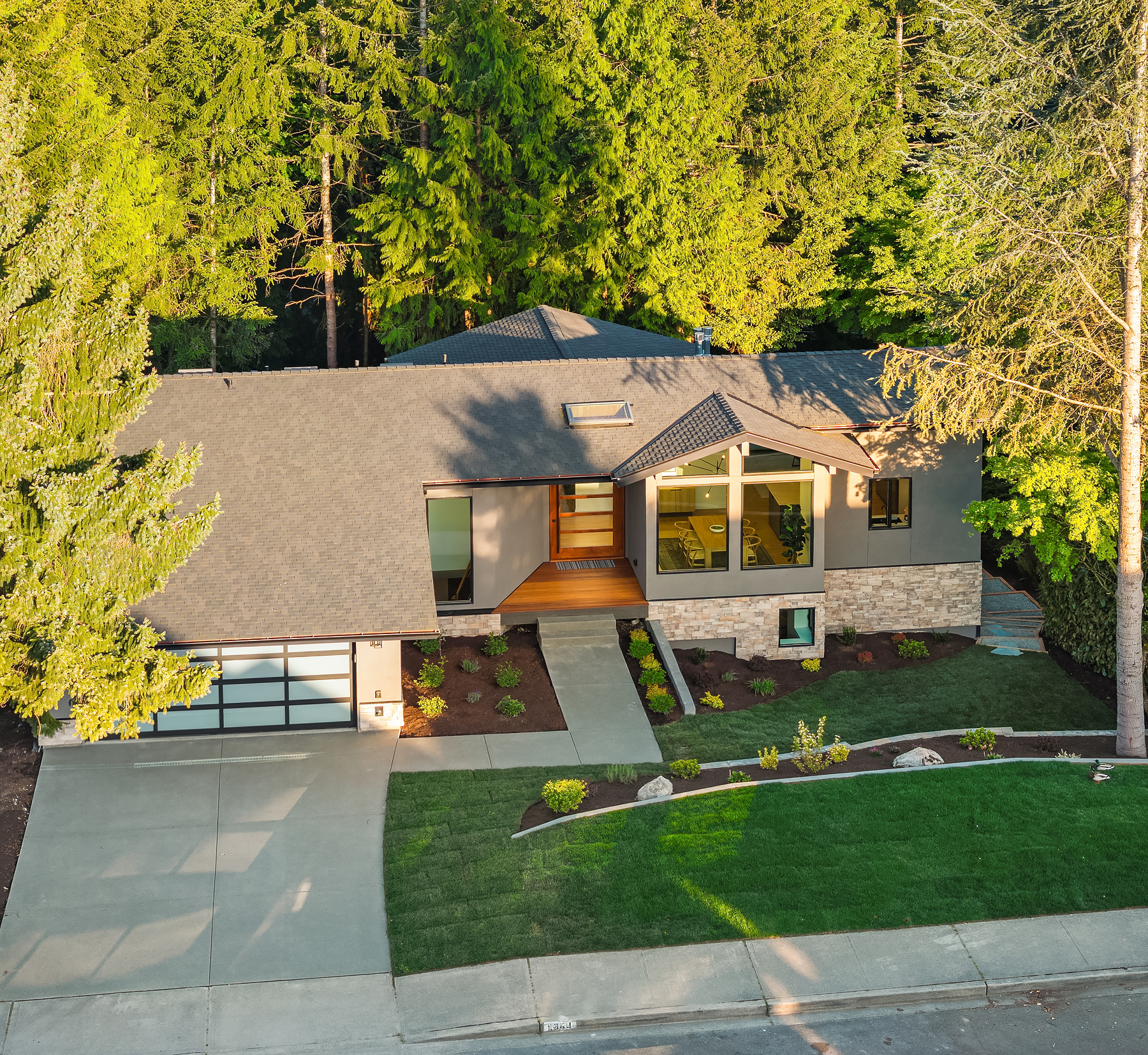 A modern single-story house with large windows, a double garage, and a neatly landscaped front yard, surrounded by tall green trees.