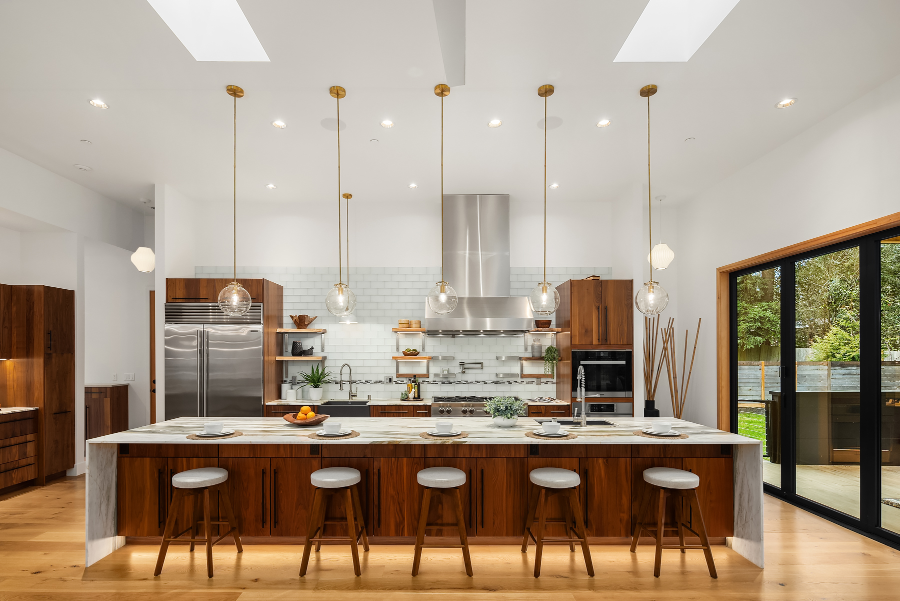 Modern kitchen with wooden cabinets, marble island with five bar stools, hanging pendant lights, stainless steel appliances, open shelves, and large sliding glass doors opening to a green outdoor area.
