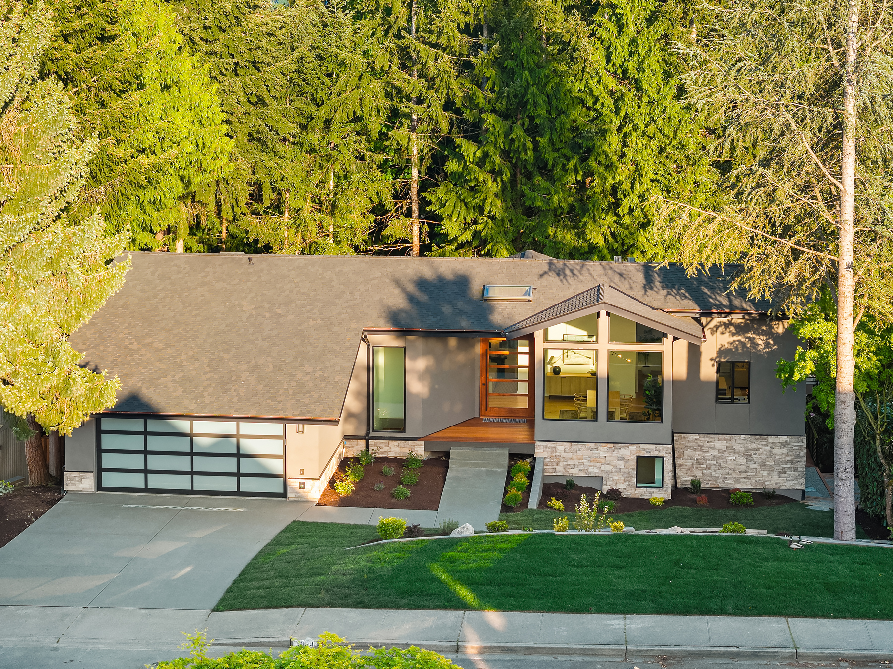 Modern single-story house with large windows, a double garage, and manicured lawn, surrounded by tall green trees. The exterior features gray walls, stone accents, and a wooden entrance porch.