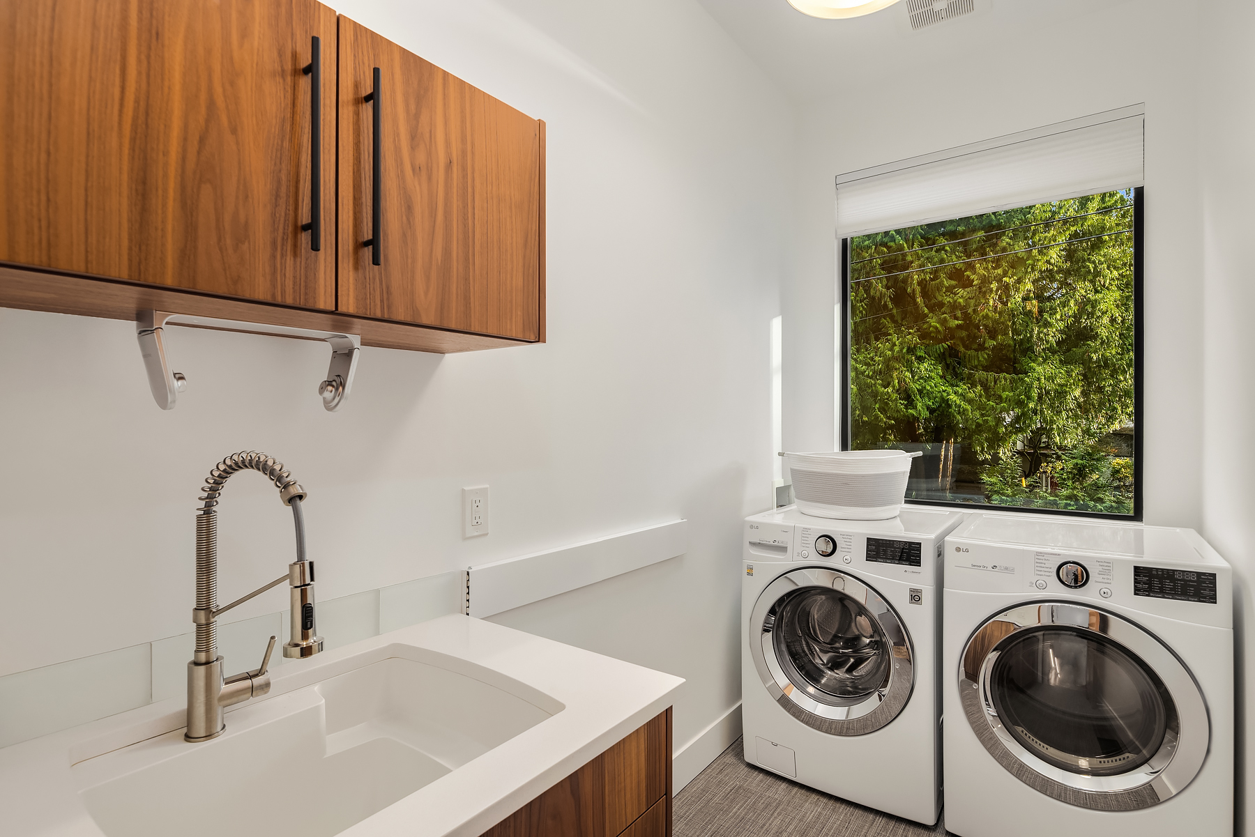 Modern laundry room with a white countertop and sink, wood cabinets, stainless steel faucet, and front-loading washer and dryer under a window with a view of green trees outside. A white laundry basket sits on the dryer.
