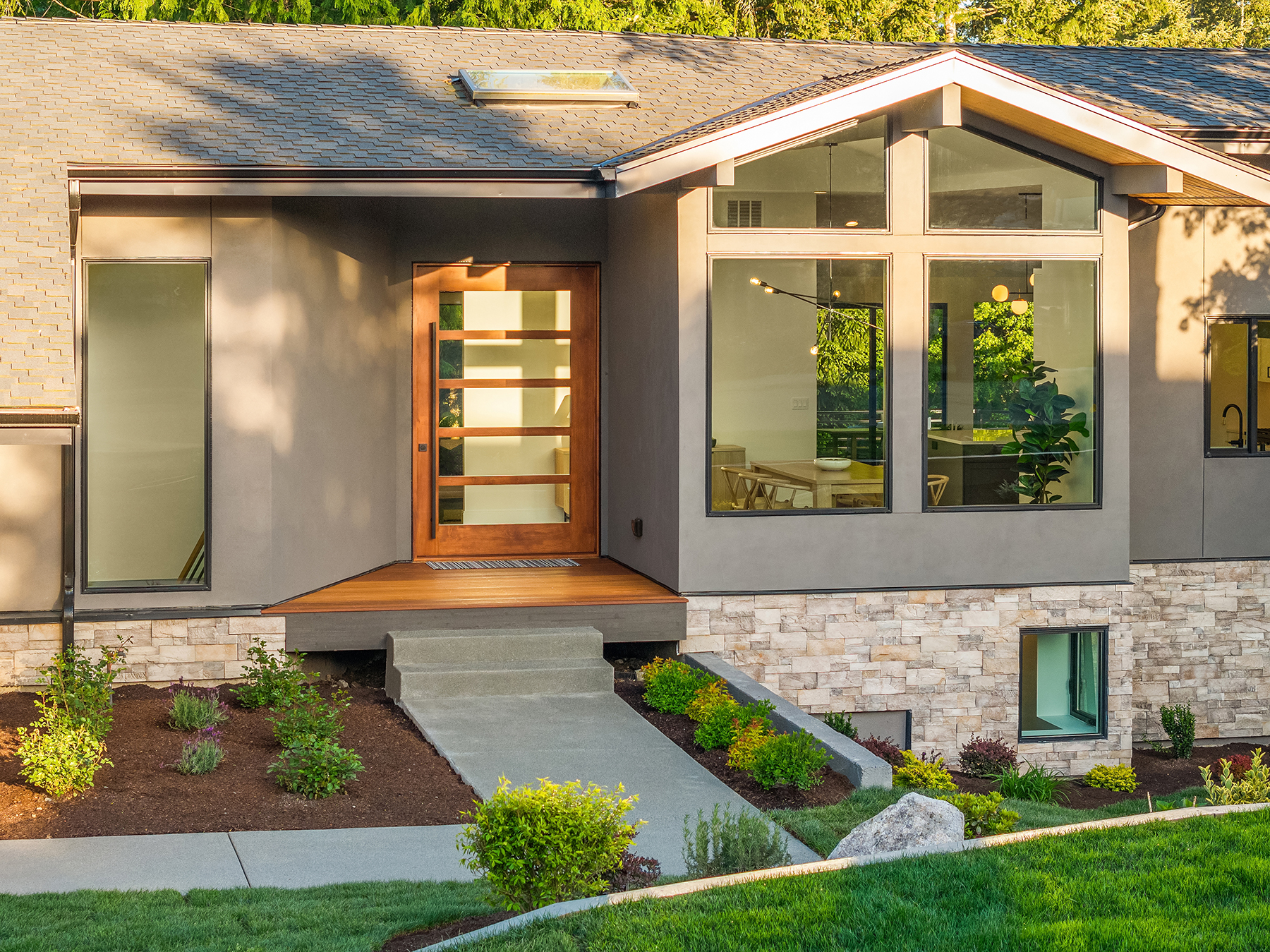 Modern house with large windows, a wooden front door with glass panels, stone accents on the lower level, landscaped yard, shrubs, and steps leading to the entrance.