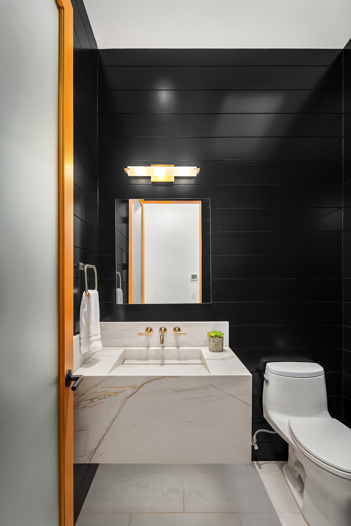 Modern bathroom with black shiplap walls, a white marble vanity with gold fixtures, a rectangular mirror, wall-mounted light, white toilet, towel ring with white towel, and a small green plant on the counter.