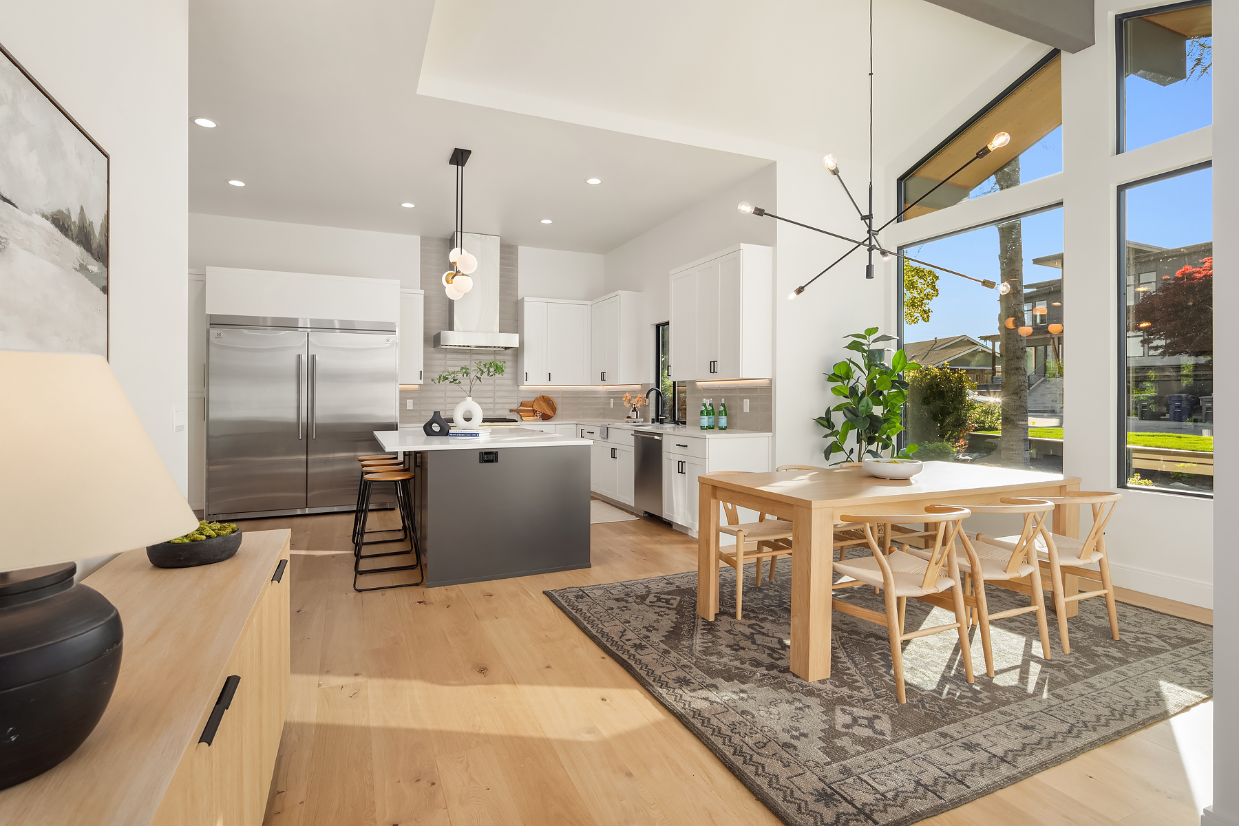 Modern kitchen and dining area with light wood floors, white cabinets, a gray island, large windows, indoor plants, and contemporary lighting, filled with natural sunlight.
