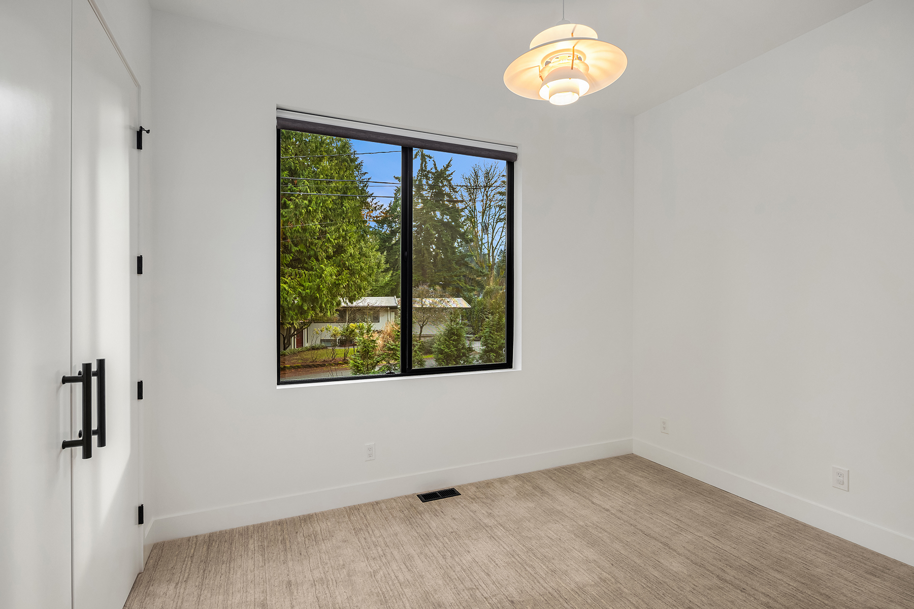 Minimalist empty room with beige carpet, white walls, a black-framed window showing trees outside, and a modern overhead light fixture. Double white doors with black handles are on the left wall.