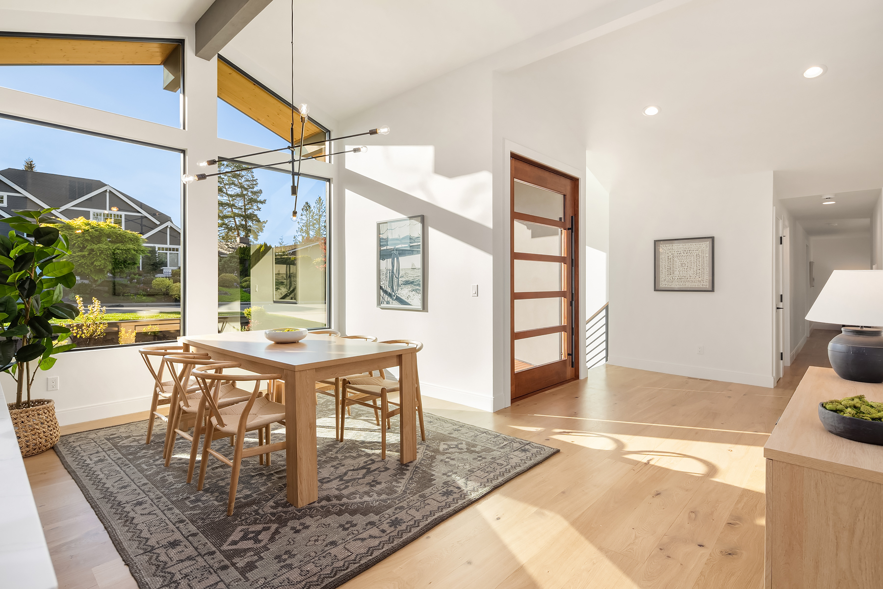 Bright dining area with large windows, wooden table and chairs, patterned rug, modern light fixture, potted plant, and view of a green neighborhood through the windows. Sunlight fills the space.