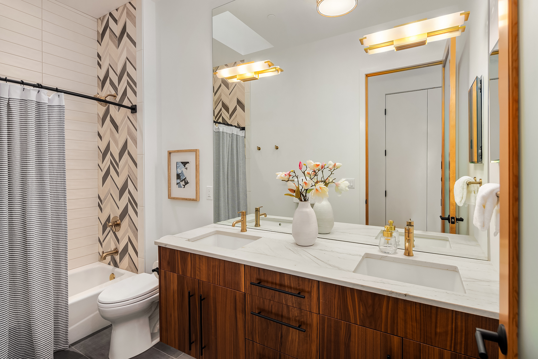 Modern bathroom with a double sink vanity, white marble countertop, wood cabinets, gold fixtures, a vase of flowers, and a bathtub with a striped shower curtain. Chevron tile accent wall behind the tub.