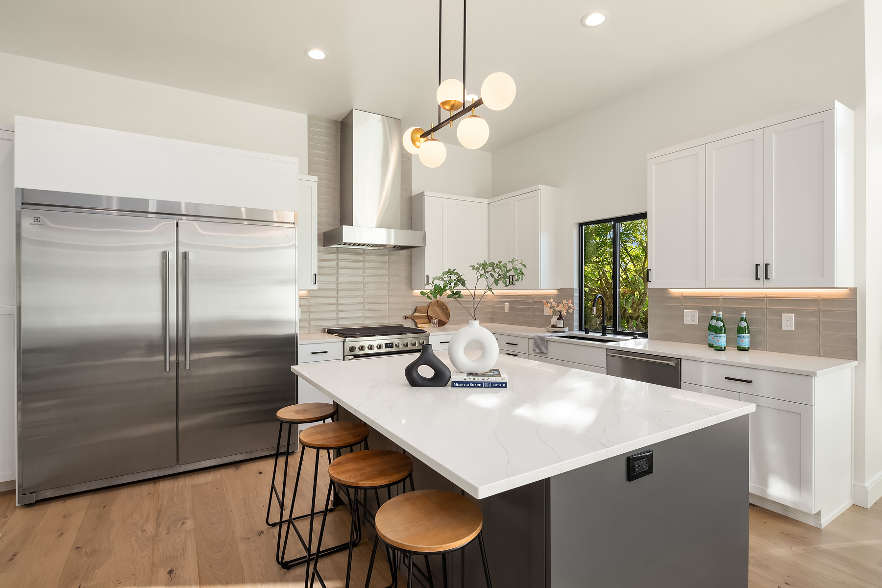 Modern kitchen with white cabinets, stainless steel appliances, a large double-door fridge, an island with a white countertop, wooden stools, and decorative objects. Pendant lights hang above the island.