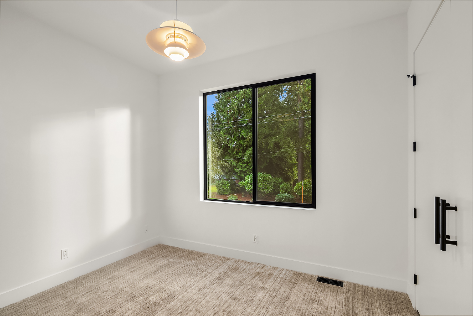 A bright, empty room with white walls, beige carpet, a modern ceiling light, a large black-framed window showing green trees outside, and a white door with black hardware.