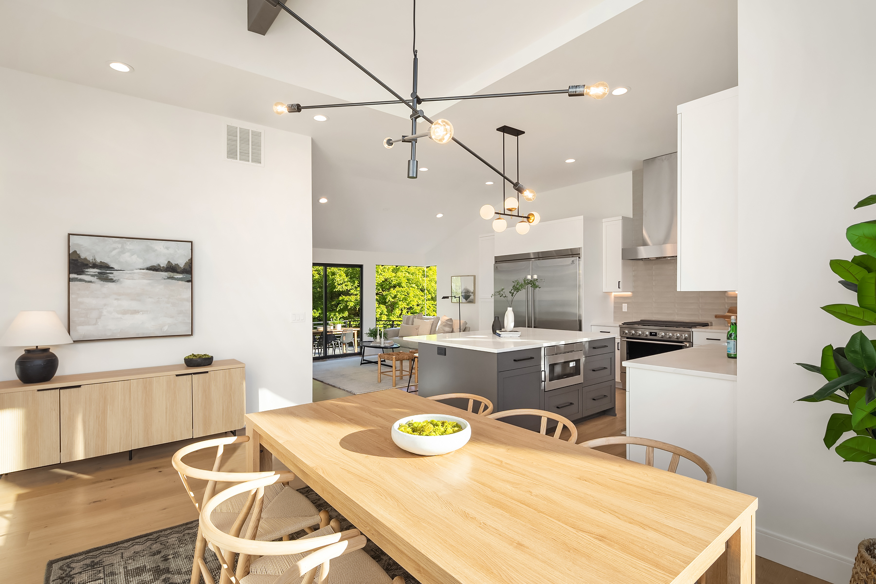 Modern dining and kitchen area with light wood furniture, a bowl of green apples on the table, minimalist decor, large windows with a view of greenery, and stylish lighting fixtures overhead.