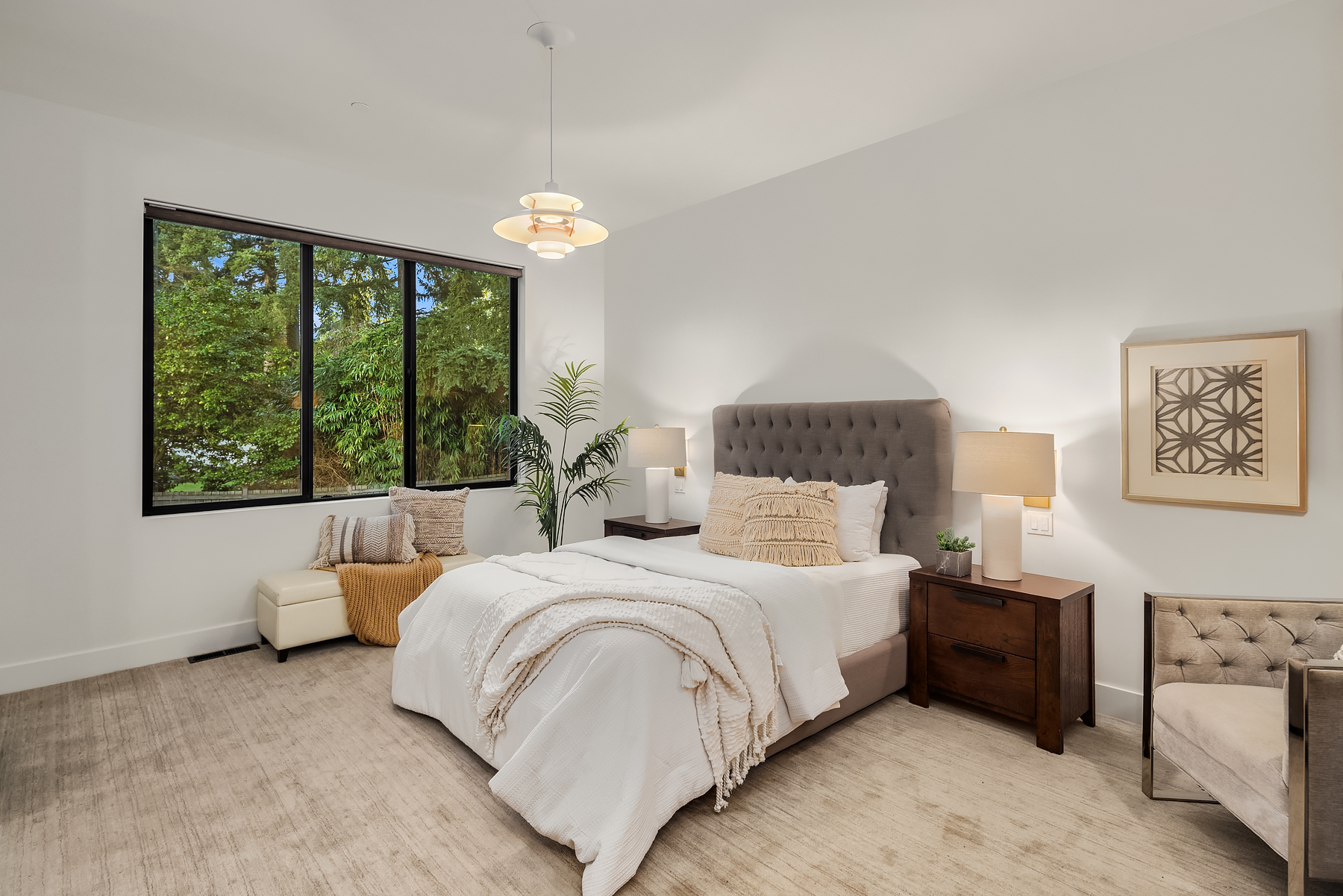 A modern bedroom with a plush bed, white bedding, two wooden nightstands, soft lighting, a large window with a leafy view, and a cozy reading nook with a chair and plant.