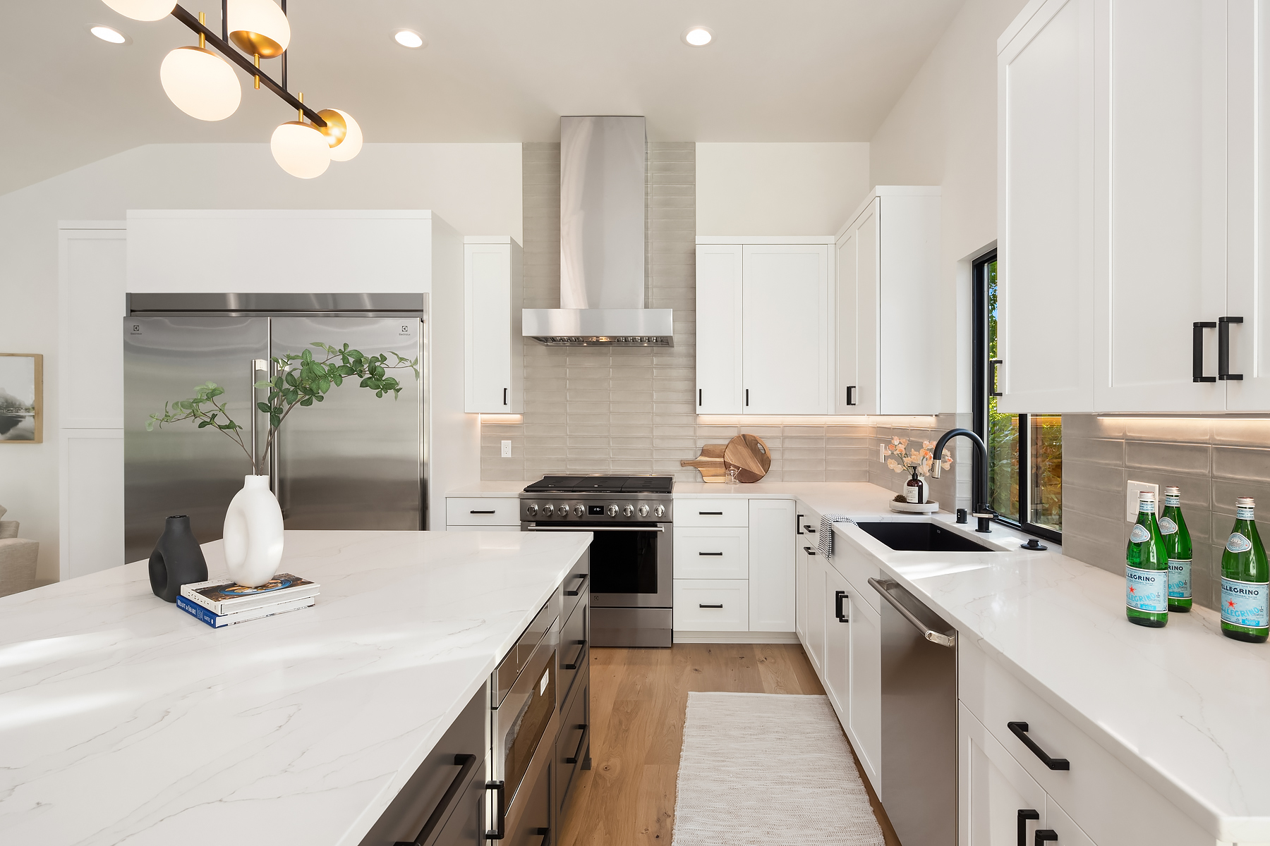 Modern white kitchen with stainless steel appliances, a large marble island, white cabinets, a farmhouse sink, and decorative items including vases and bottled water, with natural light from a window.