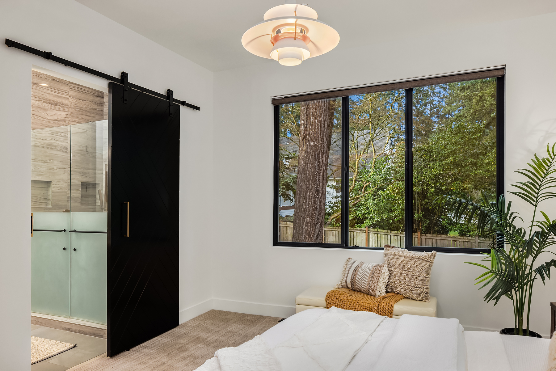 Modern bedroom with a large window showing trees outside, a black sliding barn door leading to a bathroom, a white bench with cushions and a throw, and a potted plant next to the bed.
