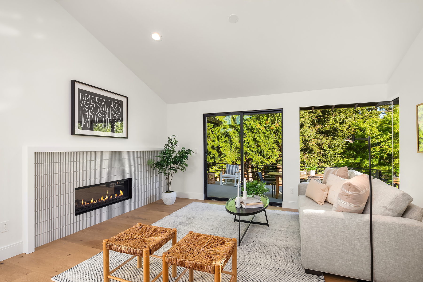 Modern living room with white walls, a gas fireplace, light wooden floors, a gray sofa with pink pillows, a round coffee table, two woven stools, a potted plant, and large windows showing green trees outside.