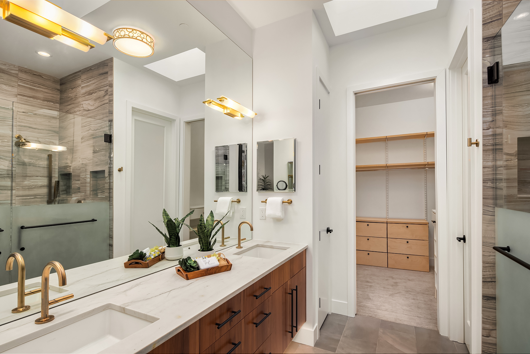 Modern bathroom with double sinks, gold fixtures, large mirror, and tray decor. A glass shower is on the left, and a walk-in closet with shelves and drawers is visible through an open door on the right.