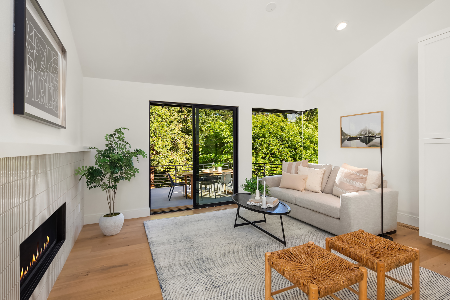 Modern living room with a light gray sofa, coffee table, two woven stools, a fireplace, a potted plant, and large glass doors leading to a balcony with lush green trees visible outside.