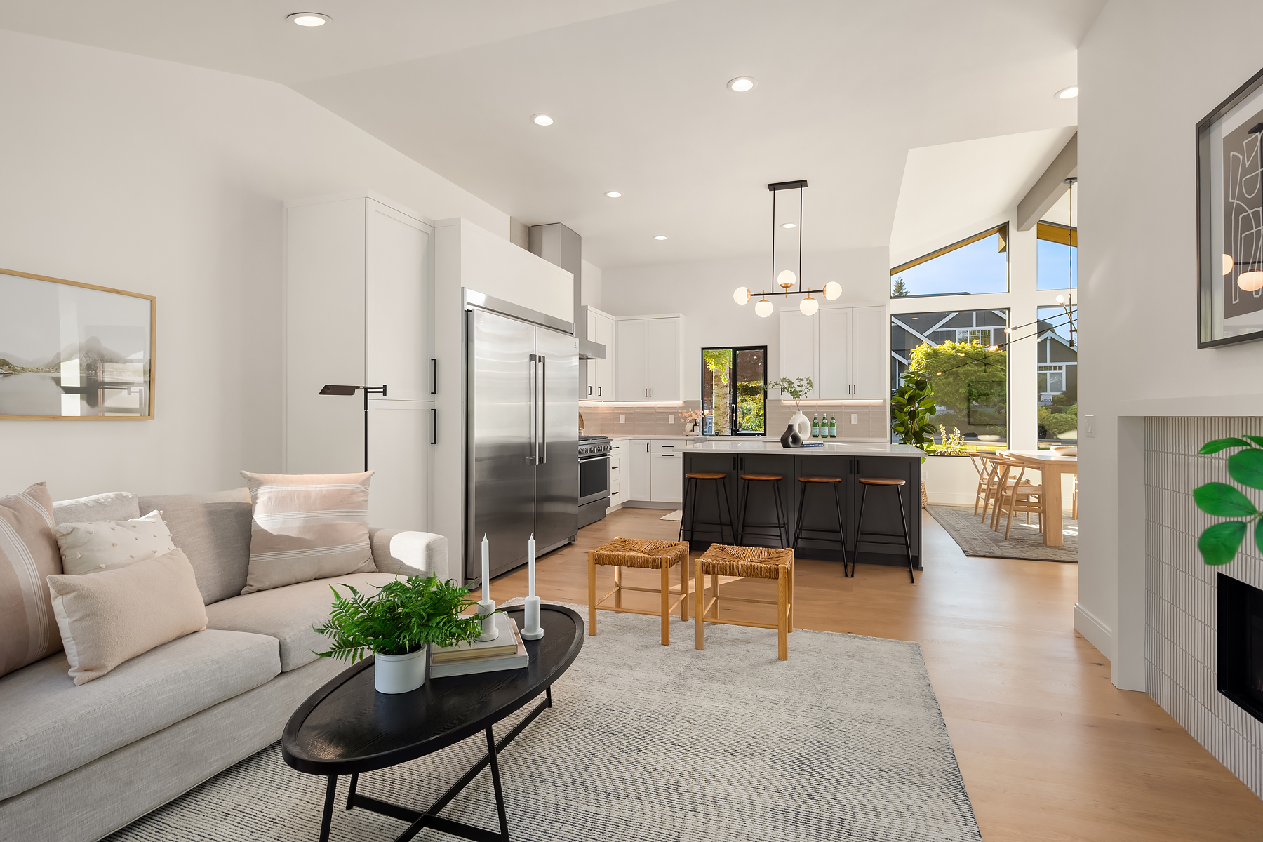 Modern open-concept living room and kitchen with light wood floors, white walls, a gray sectional sofa, black coffee table, island with stools, stainless steel appliances, and large windows letting in natural light.