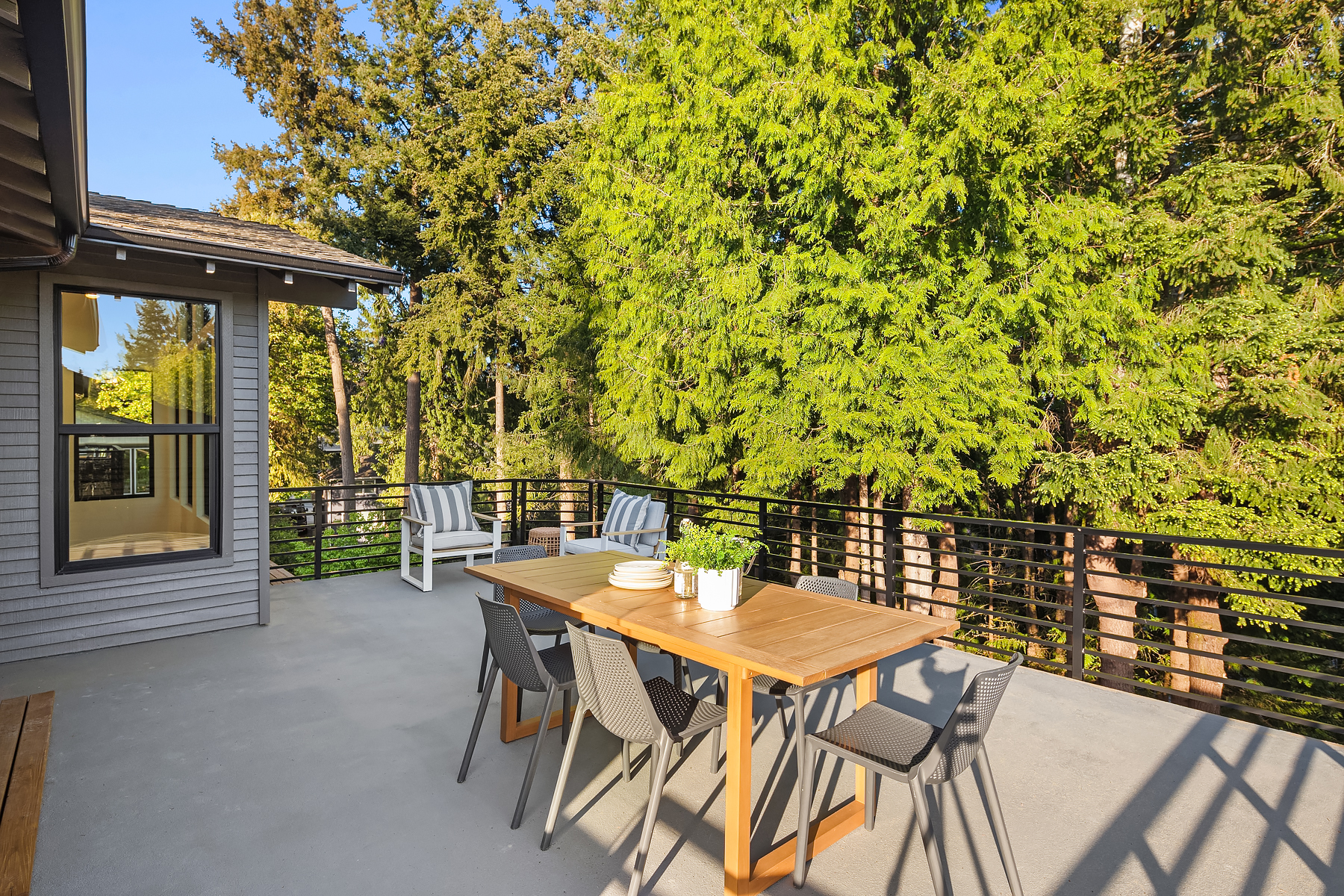 A wooden outdoor dining table with chairs and potted plants sits on a spacious patio, surrounded by a black metal railing and lush green trees. Two white armchairs are placed near the house.