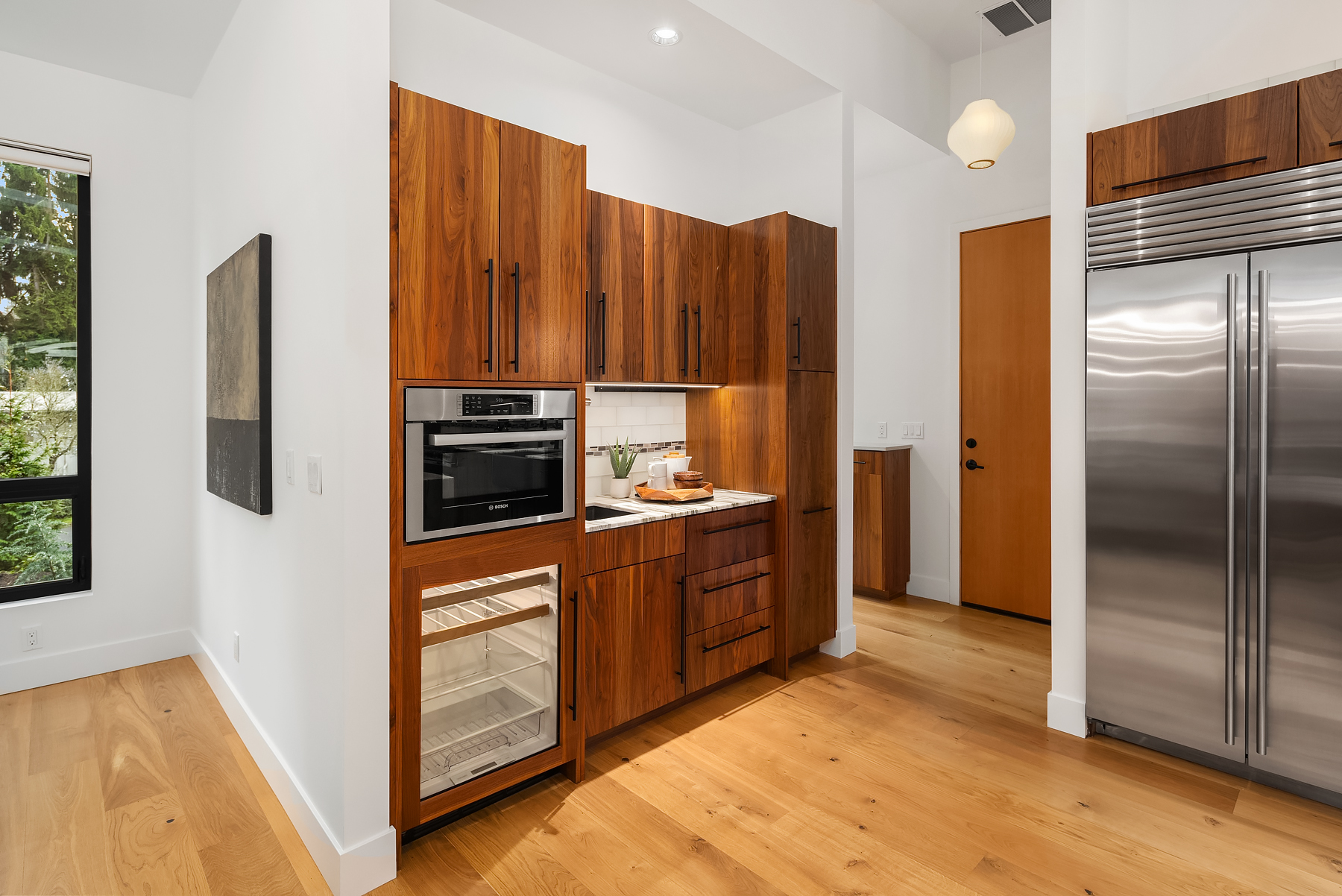 Modern kitchen with wood cabinets, built-in oven, glass-front mini fridge, and stainless steel refrigerator. Light wood floors, white walls, and a large window provide a bright, clean atmosphere.