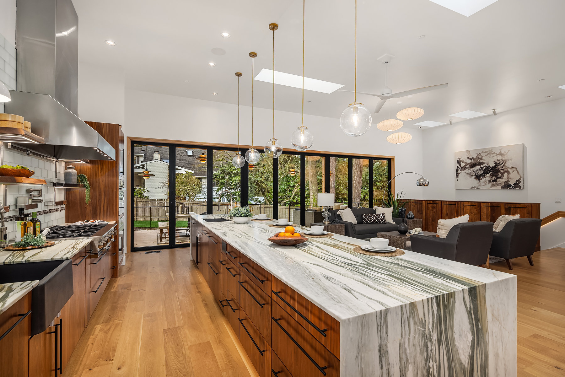 Modern kitchen with a large marble island, wooden cabinets, pendant lights, and an open layout leading to a living area with sofas and a painting. Glass doors open to a patio with greenery outside.