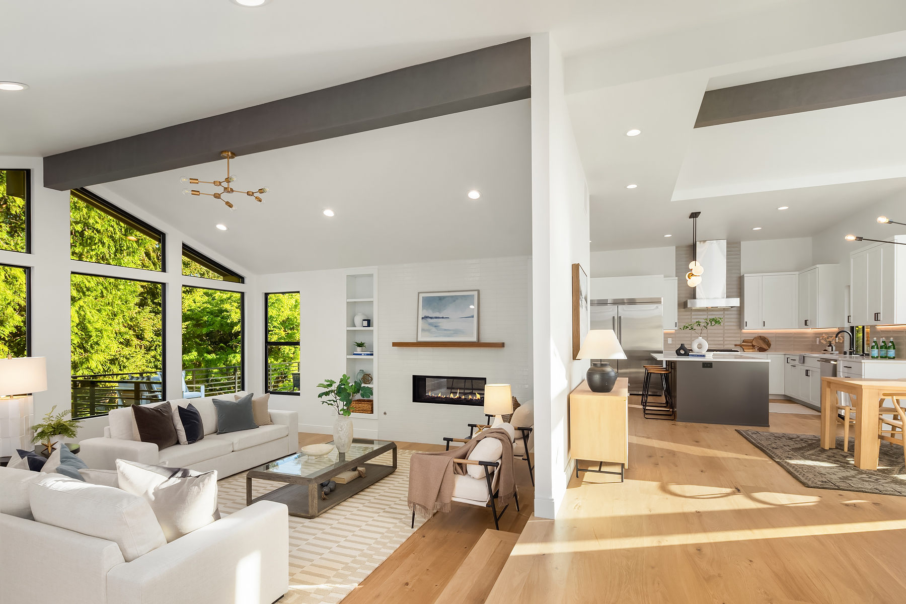 Spacious modern living area with large windows, neutral-toned furniture, a fireplace, open shelving, and a bright kitchen with white cabinets, stainless steel appliances, and a central island. Natural light fills the space.