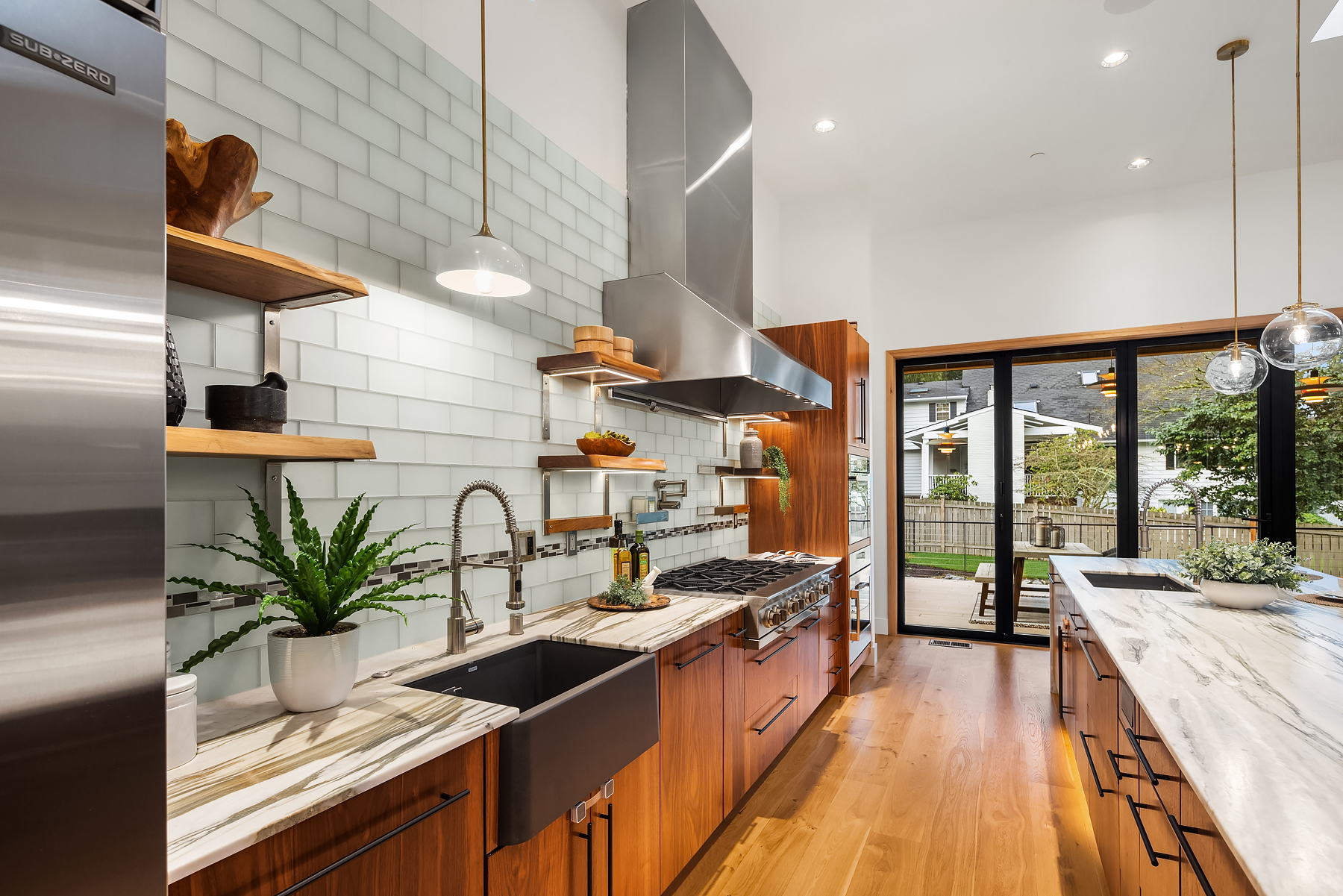 Modern kitchen with wooden cabinets, marble countertops, stainless steel appliances, a farmhouse sink, open shelves with plants and decor, glass doors leading to a backyard, and bright natural lighting.