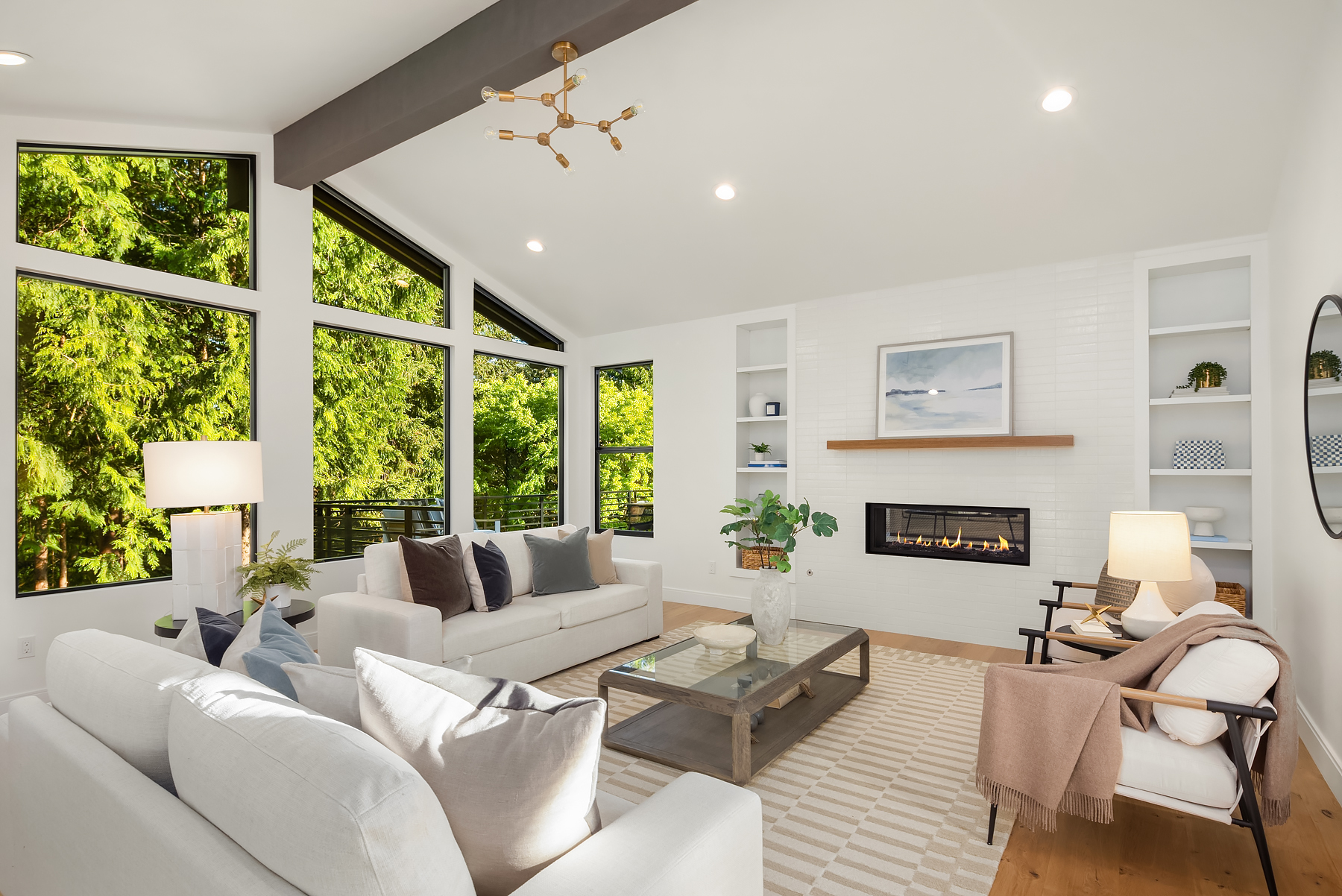 Bright modern living room with large windows, white walls, two sofas, armchairs, a coffee table, a rug, built-in shelves, a fireplace, and views of lush green trees outside.
