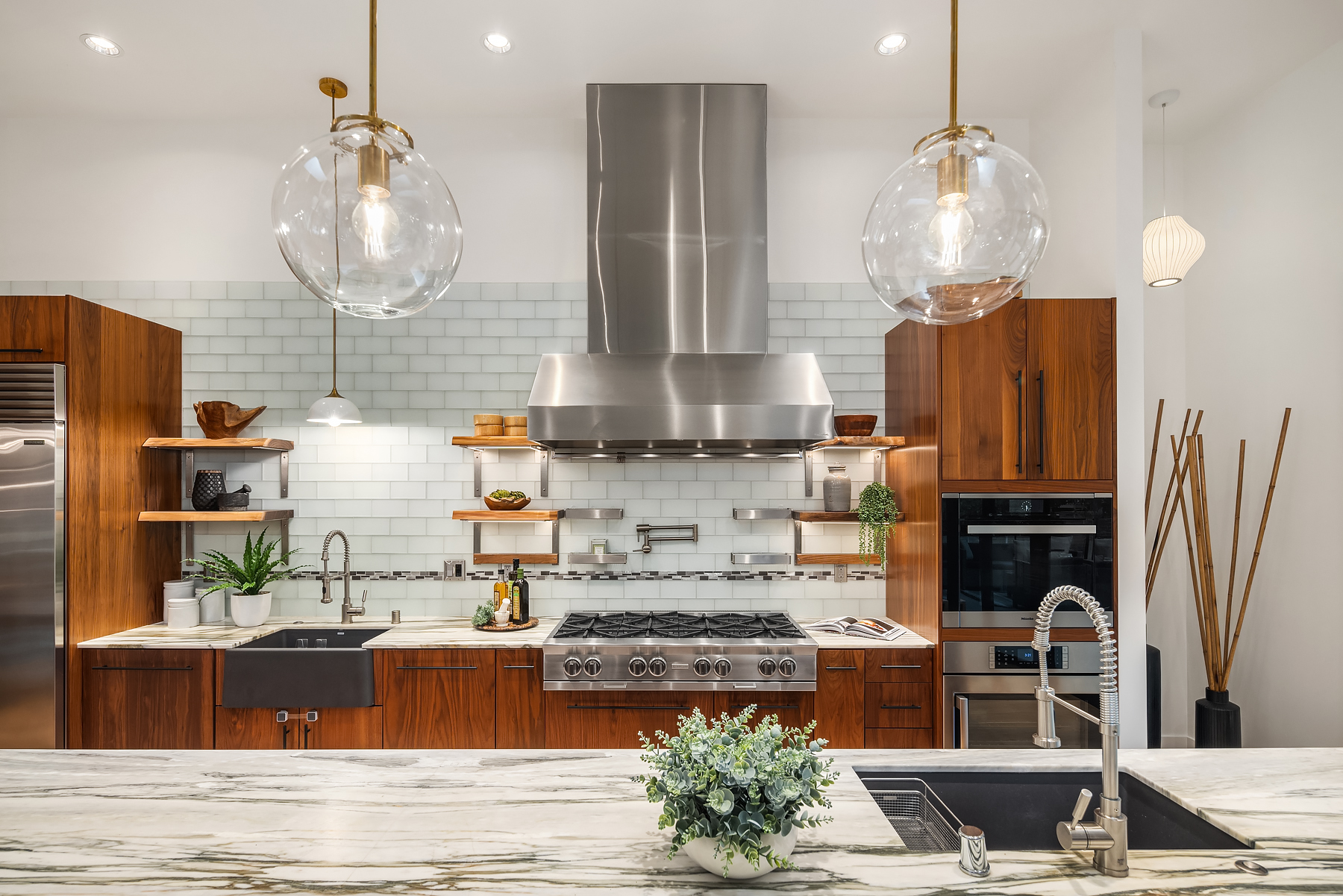 Modern kitchen with wooden cabinets, stainless steel appliances, marble countertops, a farmhouse sink, glass pendant lights, open shelves with plants and decor, and a white subway tile backsplash.