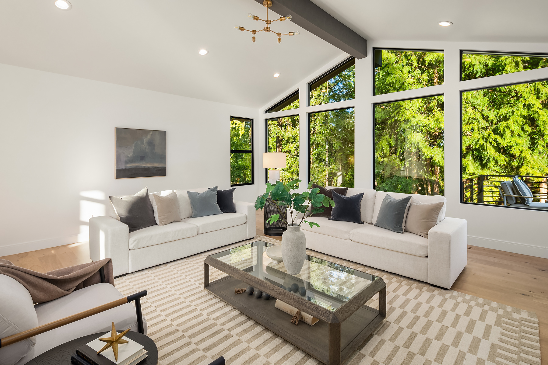 Bright modern living room with large floor-to-ceiling windows, two white sofas, a glass coffee table with a vase and decor, neutral rug, and lush green forest view outside.