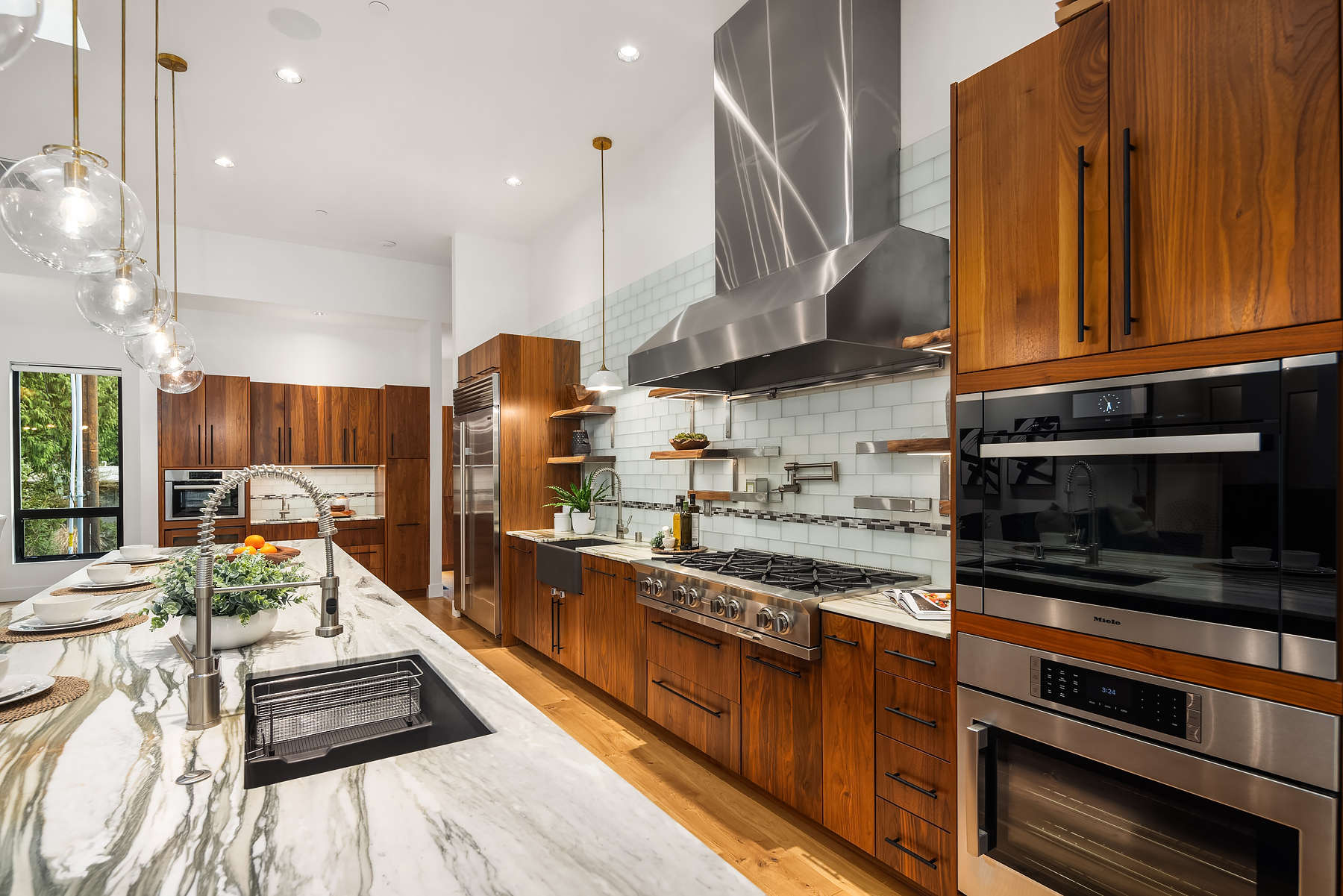 Modern kitchen with wooden cabinets, marble island with sink, stainless steel appliances, a large range hood, hanging globe lights, and bar stools. White subway tile backsplash and plants add a fresh touch.