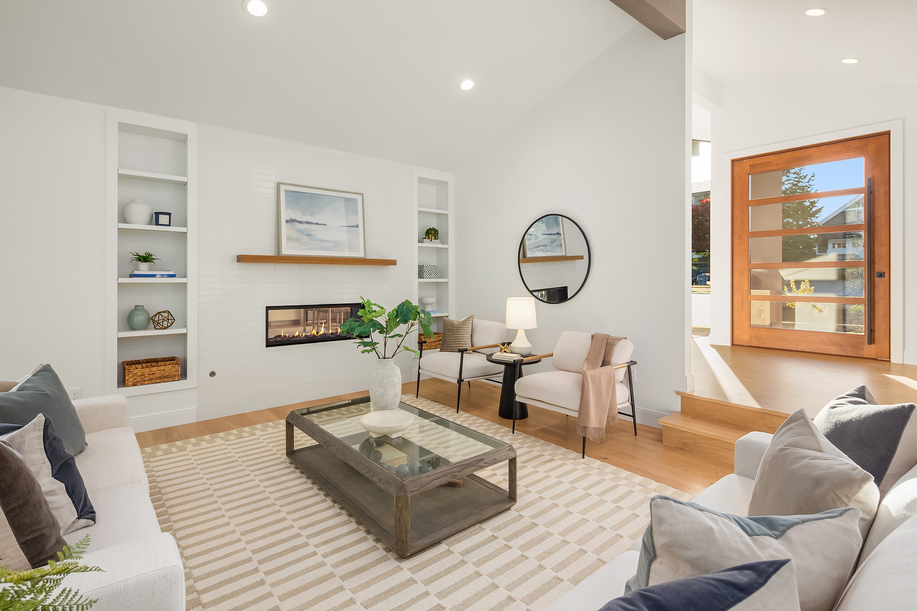 A bright, modern living room with white walls, light wood floors, a striped rug, two sofas, armchairs, a glass coffee table, built-in shelves, a fireplace, and a round mirror near a wooden front door.