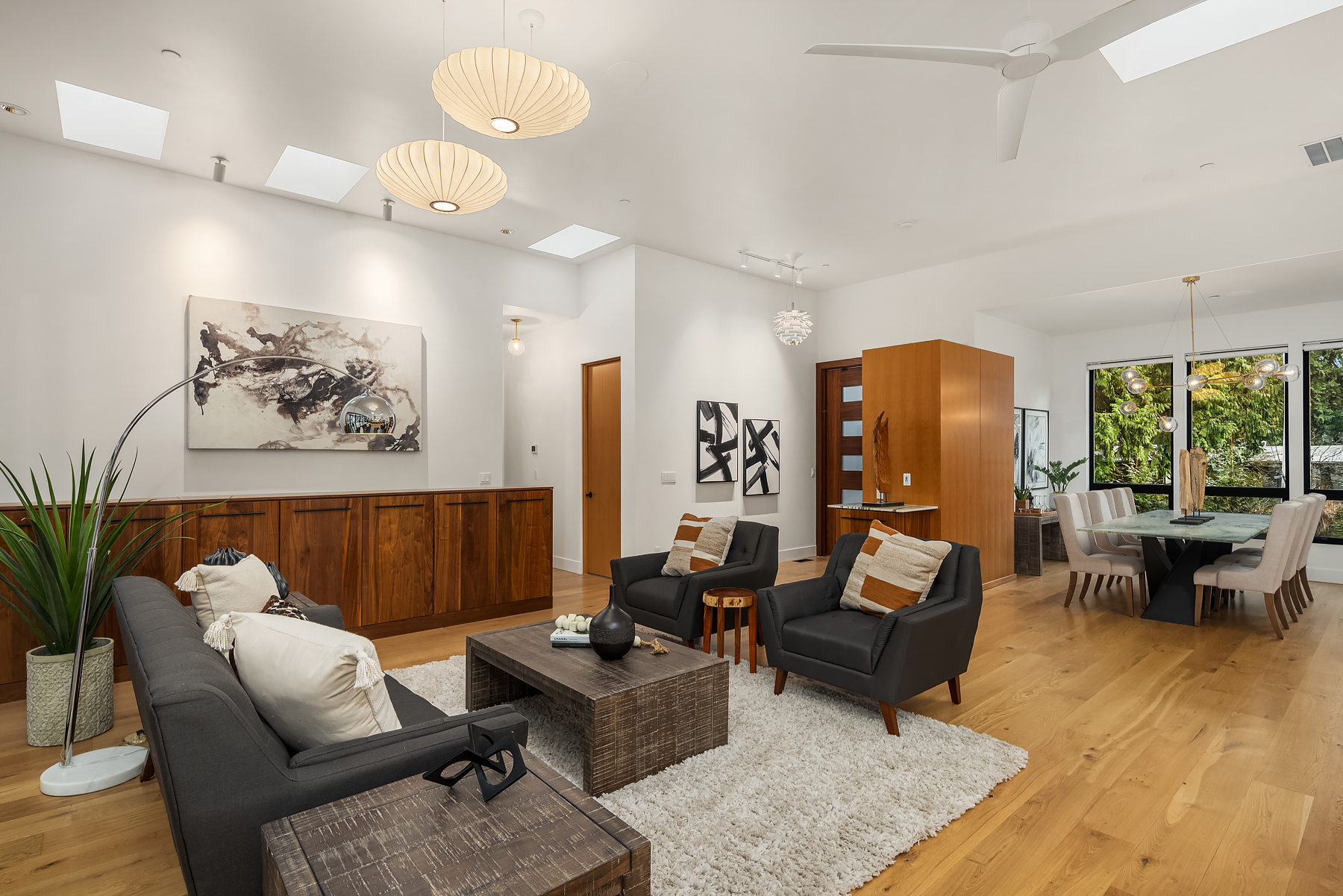 Modern living room with wooden floors, a gray sofa, two black armchairs, a coffee table, potted plants, wall art, and pendant lights; dining area visible in the background with large windows and greenery outside.
