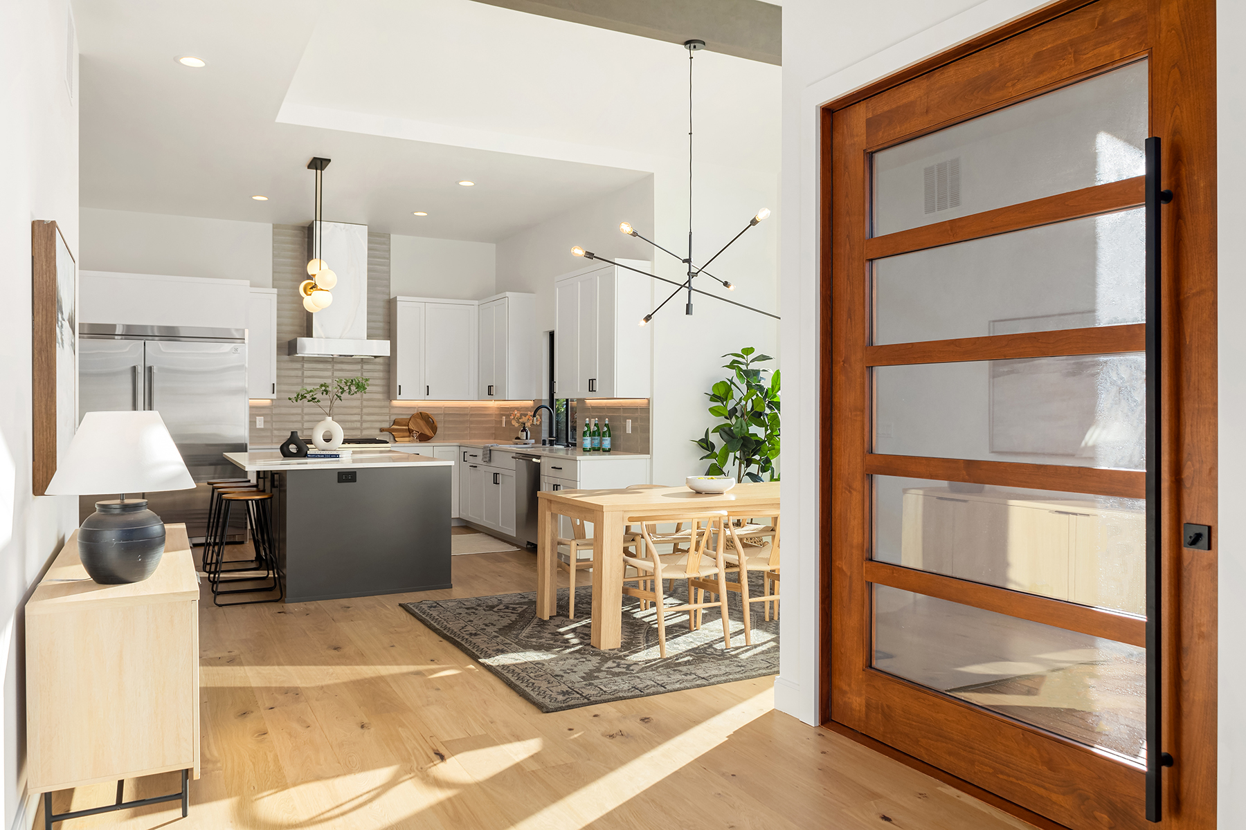 Modern kitchen and dining area with light wood floors, white cabinets, stainless steel appliances, island with gray base, wooden dining table with chairs, contemporary pendant lights, and large wooden-framed glass door.