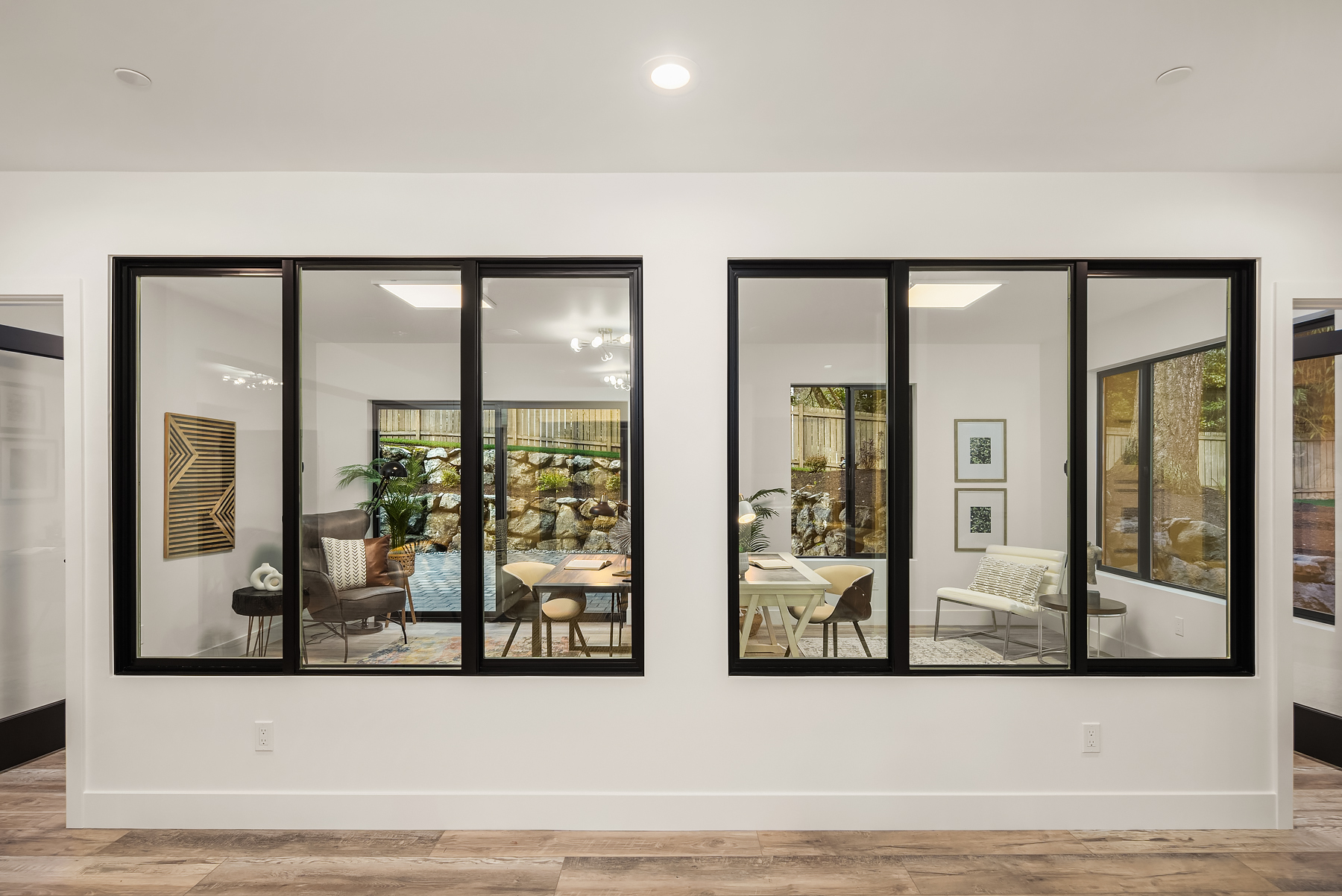 Modern interior with large black-framed glass windows dividing two office or sitting areas, featuring contemporary furniture, wall art, plants, and a view of an outdoor rock garden through more windows.